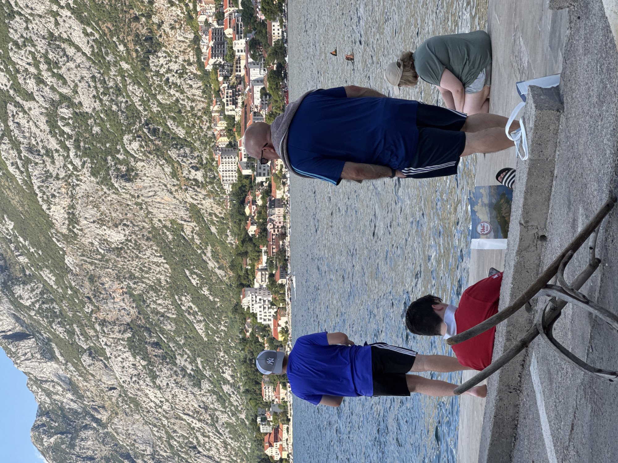 In Prčanj, Montenegro, four individuals are positioned on a concrete waterfront promenade. The focal point is a body of water, likely the Bay of Kotor, with a mountainous terrain densely covered in green vegetation and rocky outcrops rising behind a coastal town. The town features numerous buildings with red-tiled roofs and white facades, nestled along the shoreline.
On the promenade, the furthest person from the viewer, a male with a shaved head, wears a blue t-shirt with three stripes on the sleeves, dark shorts with white stripes, and sunglasses. A grey towel is draped over his shoulders. To his left, a younger male, facing away from the viewer, is dressed in a blue t-shirt and black shorts with white stripes. He wears a gray baseball cap with a white logo. In front of him, another young male is seated, facing left, wearing a red shirt and holding a mobile phone. To the right of the seated male, a female wearing a green t-shirt and light-colored shorts is also seated.
Objects on the promenade include two shopping bags near the seated individuals. One bag displays a landscape painting and text, possibly indicating it's from a local establishment. A pair of white flip-flops rests near the right edge of the frame. In the foreground, a weathered, rustic structure made of branches is positioned on the concrete. The water in the bay appears calm, with several buoys visible, and a couple of individuals can be seen swimming in the distance. The overall scene suggests a relaxed atmosphere by the water, with the backdrop of the imposing mountains and the picturesque town.