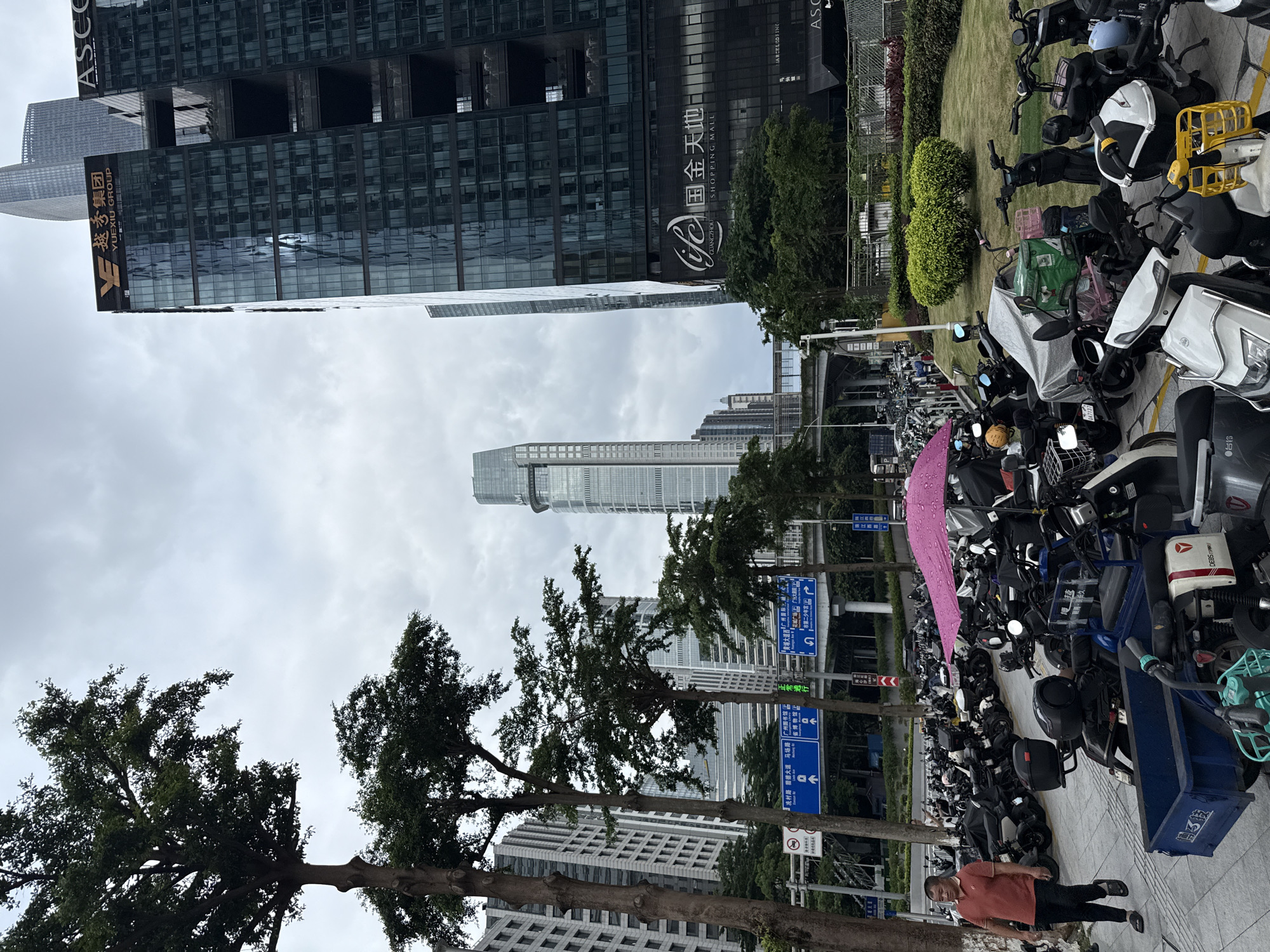 In Tianhe District, China, a man in a red polo shirt and black pants walks on a paved sidewalk next to a large parking lot filled with numerous electric scooters and motorcycles. A bright pink umbrella is open over some of the parked vehicles. In the background, modern skyscrapers dominate the skyline, with one building prominently displaying the "Yuexiu Group" logo and Chinese characters. Another building features a large sign for "Guangzhou Shopping Mall". Trees line the street and sidewalk, and an overpass structure is visible in the distance. The sky is overcast, suggesting a cloudy day.