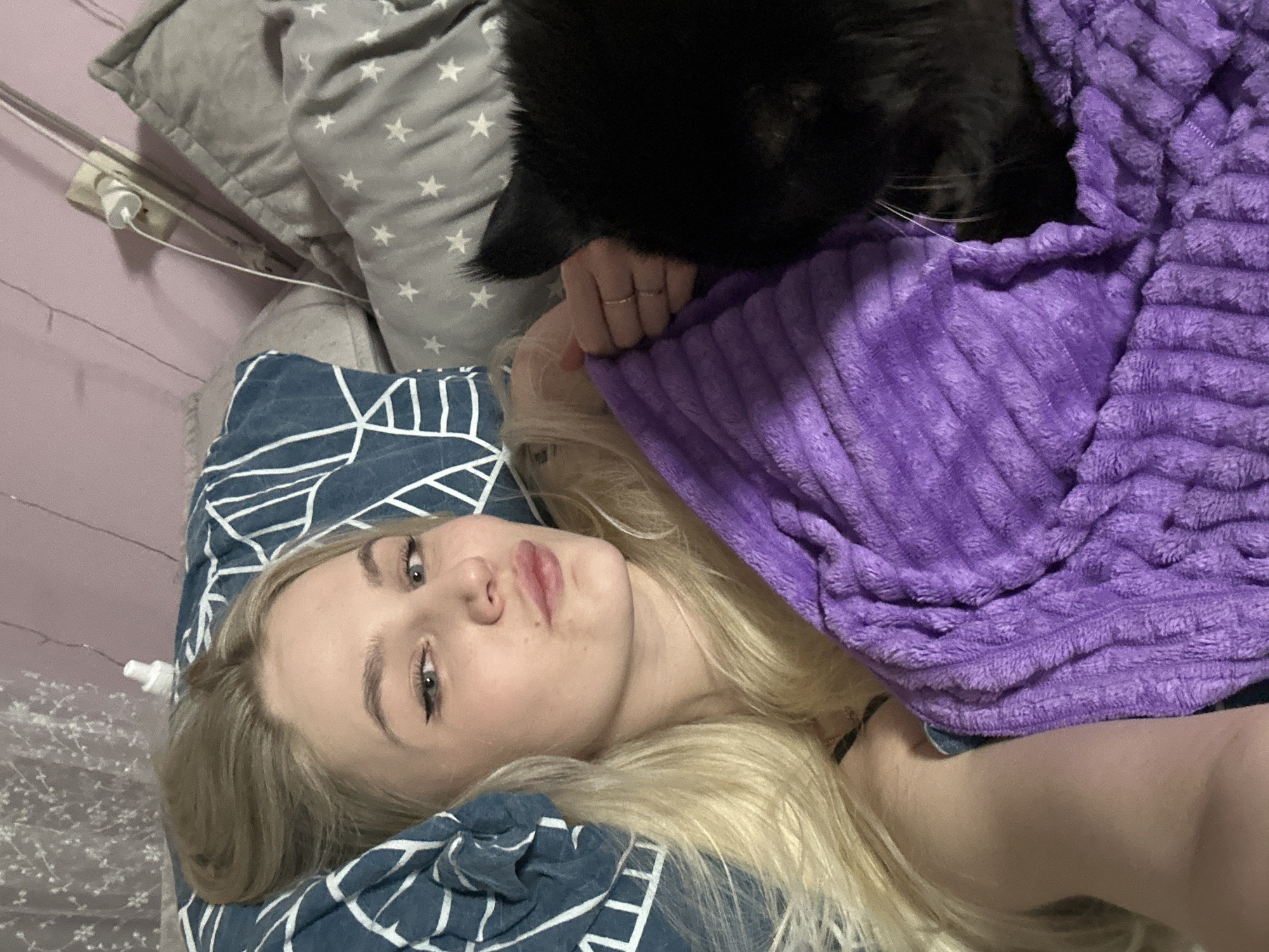 The image captures an intimate indoor scene in Chaikovsky, Russia, featuring a young woman relaxing in bed with a black cat. The woman, who has long blonde hair, lies on her back, her head resting on a blue pillow with a white geometric pattern. She looks towards the camera with a neutral to slightly relaxed expression, wearing light makeup, including eyeliner. Her left hand, wearing a ring, is visible near her shoulder, tucked under a soft, ribbed purple blanket that covers much of her upper body.
A black cat is snuggled close to her head, largely obscured by the blanket, but its dark fur and prominent white whiskers are clearly visible. The cat appears to be resting or grooming itself, suggesting a moment of quiet companionship. In the background, a light pink or lavender wall is visible, along with a grey pillow adorned with white stars. A white power outlet with a charging cable plugged in can be seen on the wall. Faint string lights are also visible in the left background, contributing to a cozy and relaxed atmosphere, likely indicating evening or low light conditions indoors. There is no visible text in the image.