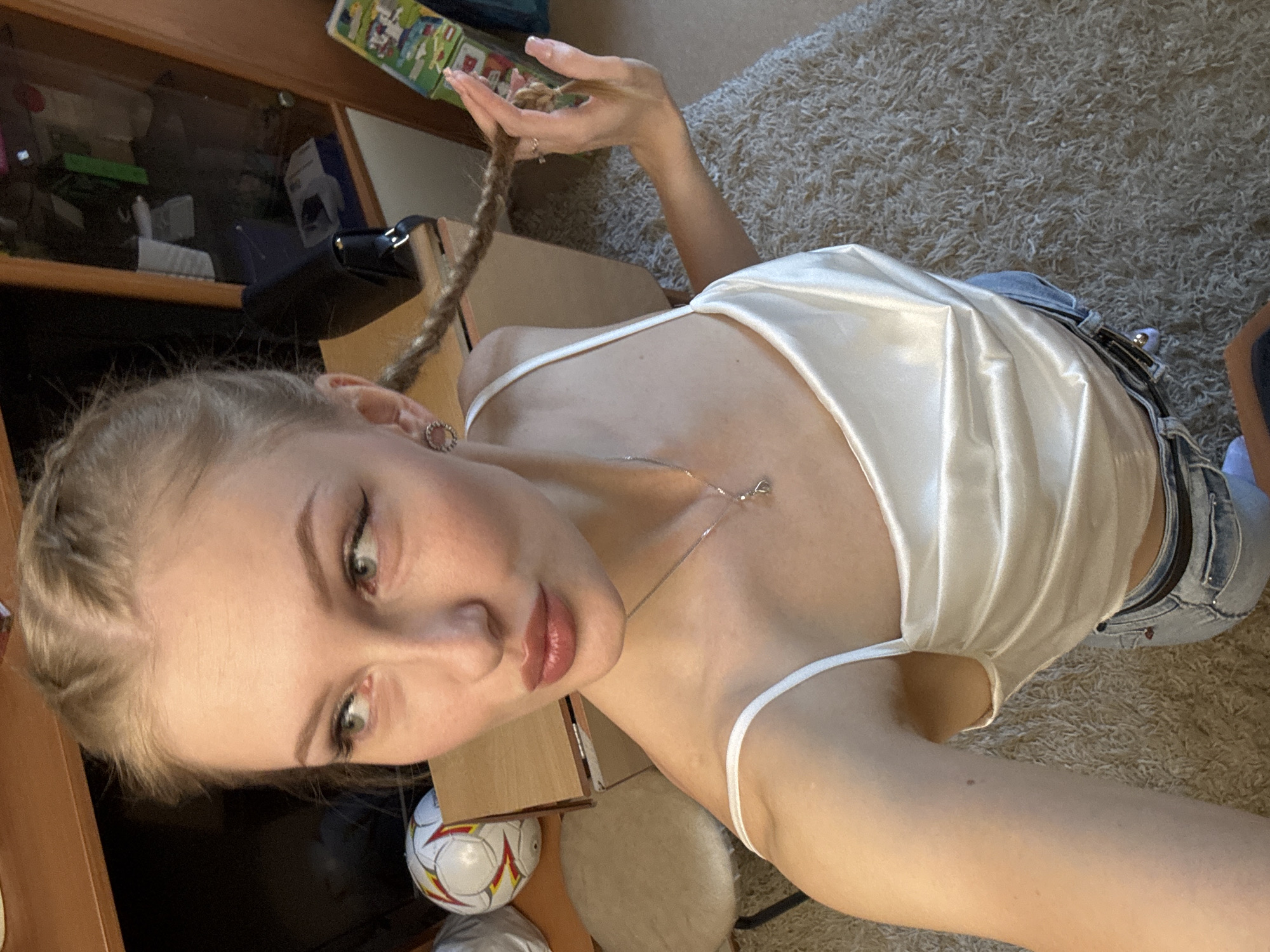 A young woman with blonde hair braided into pigtails poses for a selfie in what appears to be a home environment. She is wearing a white satin camisole and blue jeans. She is looking to her left, with her right hand touching one of her braids. In the background, there is a wooden cabinet with shelves containing various items, a brown carpet, and a small stool. A soccer ball is visible on a wooden surface to the left of the frame. The overall impression is an informal, indoor setting. There is no indication of the city of Yekaterinburg, Russia.