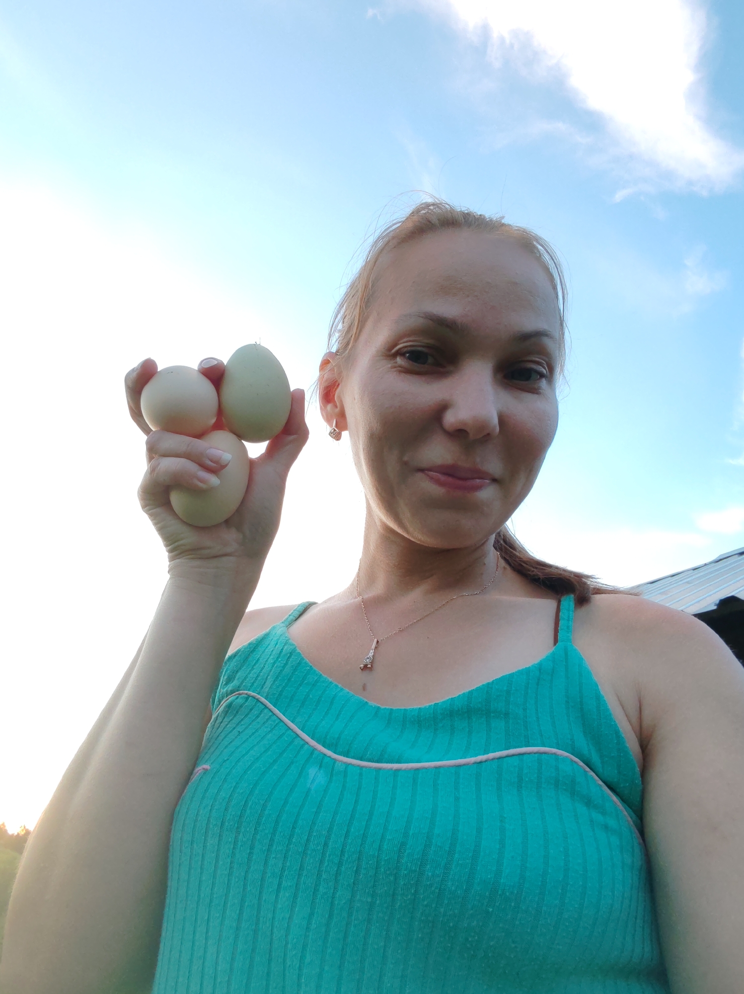 A woman is holding three eggs in her hand against a bright sky with some clouds. She is wearing a teal tank top with a thin pink trim. She is also wearing a necklace with a pendant that appears to be the Eiffel Tower. The woman is outdoors, with what looks like a building structure in the background. The image was taken in Chukhlem, Russia.