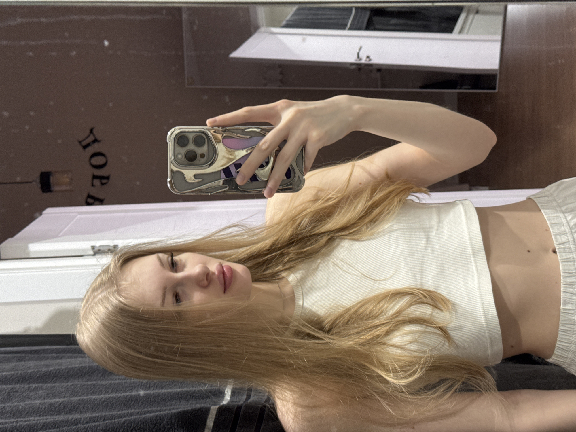 A young woman with long, blonde hair is taking a selfie in what appears to be a bathroom. She is holding a phone with a decorative case up to a mirror, which reflects the room. The background includes a brown wall with some decorative lettering, a light fixture, and a white countertop with a basin. The woman is wearing a white tank top and light-colored shorts or pants. The lower portion of the image shows what appears to be a dark-colored piece of furniture or bedding with a vertical stripe.
I have no information that allows me to confirm that the image was taken in the city of Perm, Russia.