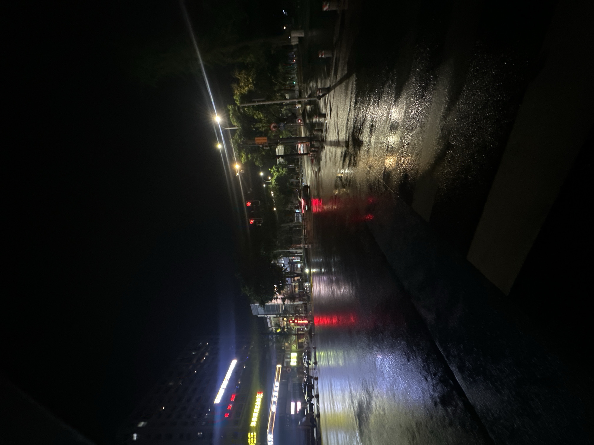 The image presents a nighttime urban street scene in the Chongqing High-tech Zone, China, characterized by a wet asphalt road surface suggesting recent rainfall. The foreground features the dark, reflective pavement, with a partial pedestrian crosswalk delineated in the bottom right. In the midground, a multi-lane road leads to an illuminated intersection. The left side displays commercial buildings with multiple brightly lit signs, predominantly featuring Chinese characters. Several vehicles, including dark passenger cars and a larger truck-like vehicle, are present on the left lanes, some appearing stationary. A red traffic light is clearly visible on the left. The right side of the midground is illuminated by intense streetlights casting glares and reflections on the wet road. Green tree foliage lines this section, accompanied by utility poles, street signs, and a series of dark bollards acting as a road divider. Another set of red traffic lights is active on the right side of the intersection. The background is indistinct, showing general urban lighting against a dark night sky. No discernible human figures are present.