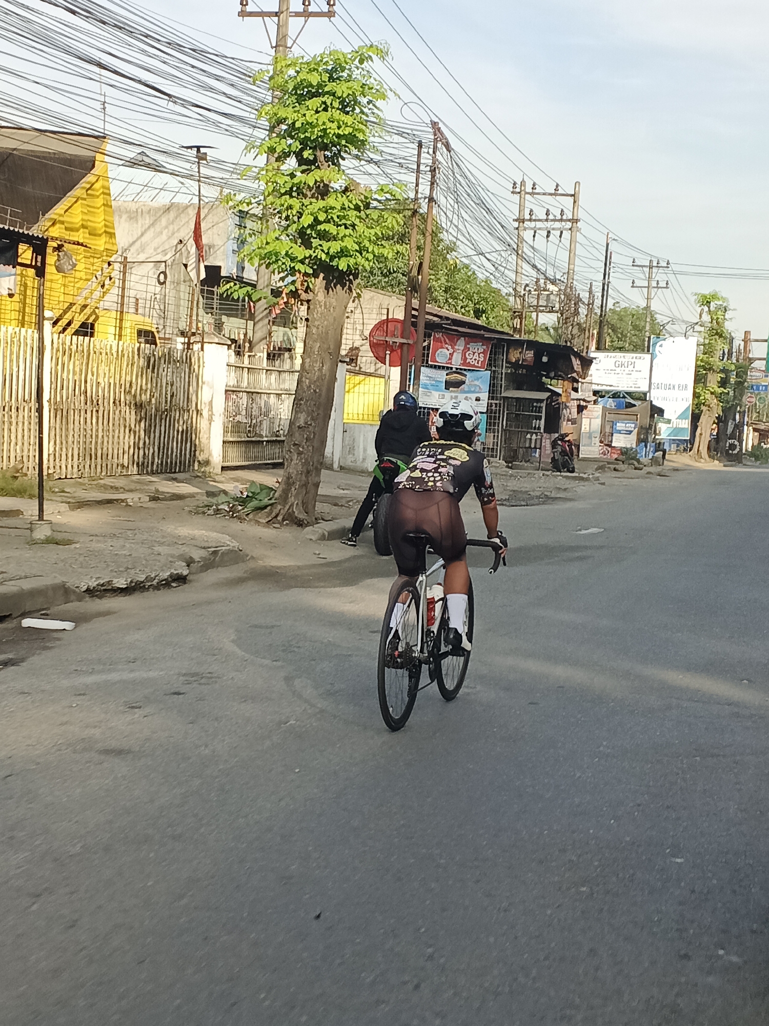 The image shows a cyclist riding a road bike on a paved street in Tg Morawa A, Indonesia. The cyclist is wearing a black cycling jersey with colorful graphics and black cycling shorts, a white helmet, and white socks. They are riding away from the camera, with their back to the viewer. The road appears to be in a semi-urban area, with buildings and power lines visible on the side of the street. A yellow building with a wooden fence is on the left side of the image, and a small shop with various signs is on the right. The sky is clear and blue, suggesting it is daytime. There is also a motorcycle parked on the side of the road near the shop. The ground surface is asphalt and there is a sidewalk area that is cracked and uneven in places.