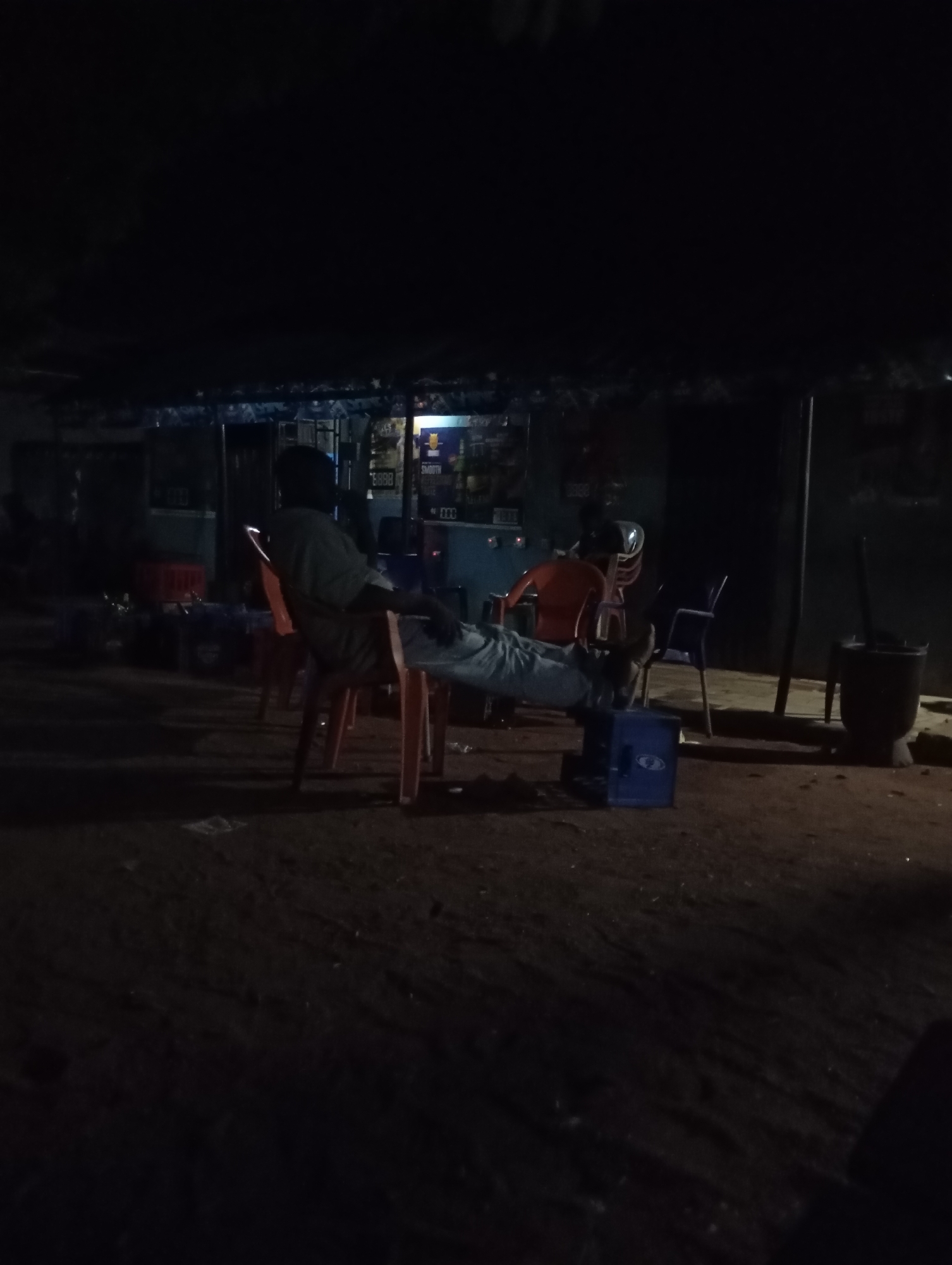 At night, in Shendam, Nigeria, a man in a light-colored t-shirt and pants is seated on an orange plastic chair. His legs are propped up on a blue plastic crate in front of him, and his arms are resting on the back of the chair. Another person, wearing an orange shirt, is seated in the background on a red chair, facing away from the camera. Behind them, a dark structure with a corrugated metal roof appears to be a shop or bar, illuminated by internal lights. Various items, including bottles and crates, are visible on shelves inside. To the right, a tall black trash bin stands near a dark pole. The ground is unpaved, appearing to be dirt or gravel. The scene suggests a casual, outdoor gathering space.