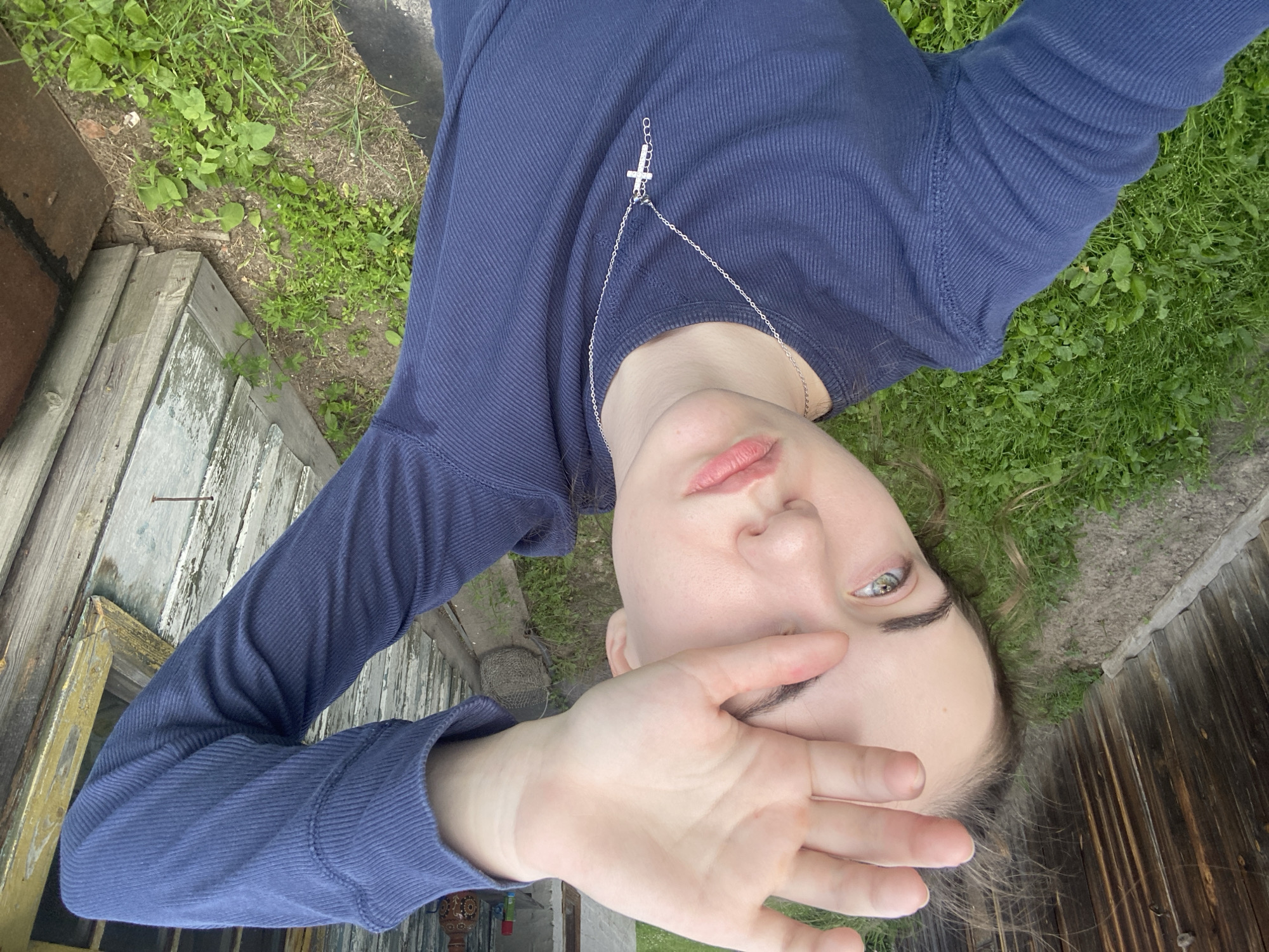 The image shows a young person lying on the ground outdoors. They are wearing a long-sleeved blue shirt with a ribbed texture and a silver cross pendant necklace. Their right hand is raised and partially covers their face, obscuring their eyes. The person has fair skin and light-colored eyes, with their lips slightly parted. They appear to be looking towards the camera.
The setting includes a patch of green grass and soil, with some scattered weeds. To the left, there is an aged wooden structure, possibly part of a fence or building, with peeling white paint. Further to the left, a section of reddish-brown brickwork is visible. To the right, a weathered wooden plank fence runs horizontally. The ground is uneven and appears to be in a somewhat rural or overgrown area.
The lighting suggests it is daytime, likely overcast or with diffused sunlight, as there are no harsh shadows. The overall mood is casual and introspective. There is no visible text in the image. The provided location context is Zavodoukovsk, Russia.