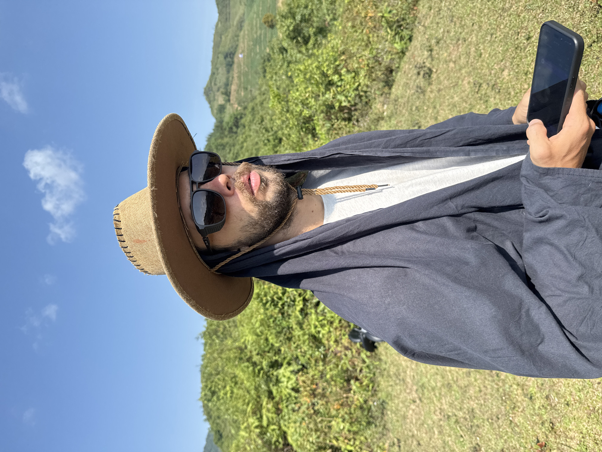 The image shows a person, likely a man, outdoors with a landscape in the background, potentially near Bản Phố, Vietnam. He is wearing a wide-brimmed hat and dark sunglasses. A dark-colored garment drapes over him. He is holding a mobile phone in his right hand. The background consists of green hills, likely with vegetation, and a clear blue sky with a few clouds. The overall lighting suggests it is daytime. The image appears to be taken from a low angle, looking upwards.