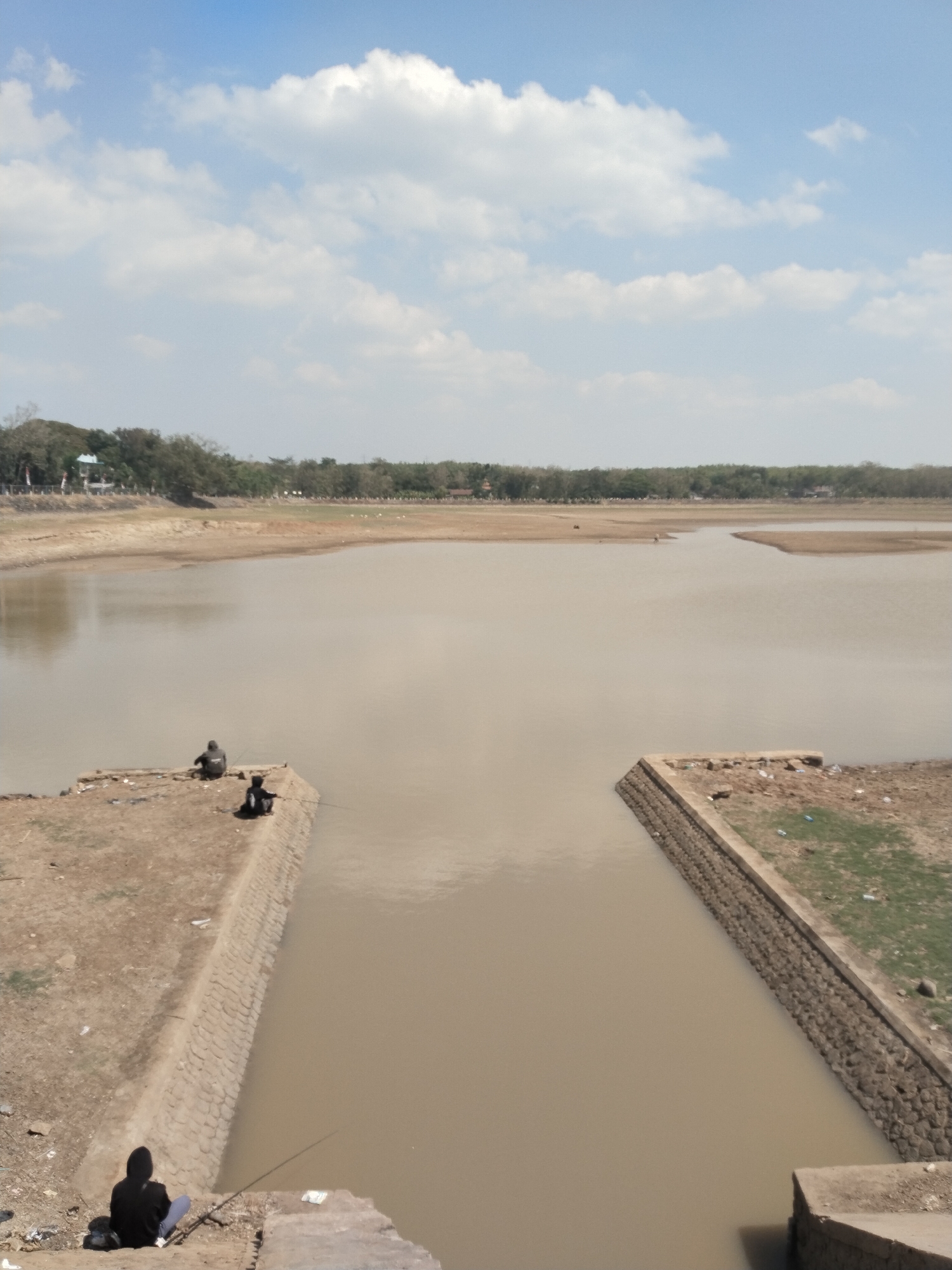 The image captures a tranquil scene at a reservoir in Blimbing, Indonesia. The sky is a vibrant blue, dotted with fluffy white clouds. The vast expanse of the reservoir dominates the frame, its murky brown water reflecting the sky. Two individuals are seen fishing by the water's edge. One person, dressed in dark clothing, sits on a concrete embankment, holding a fishing rod. Further down, another person in a black hoodie is also engaged in fishing, their rod cast into the water. The surrounding landscape is dry and barren, with sparse vegetation and a few scattered trees in the distance. The scene conveys a sense of peace and quiet, with the fishermen engrossed in their activity, undisturbed by their surroundings. The reservoir appears to be a popular spot for local anglers, offering a serene escape from the hustle and bustle of daily life.