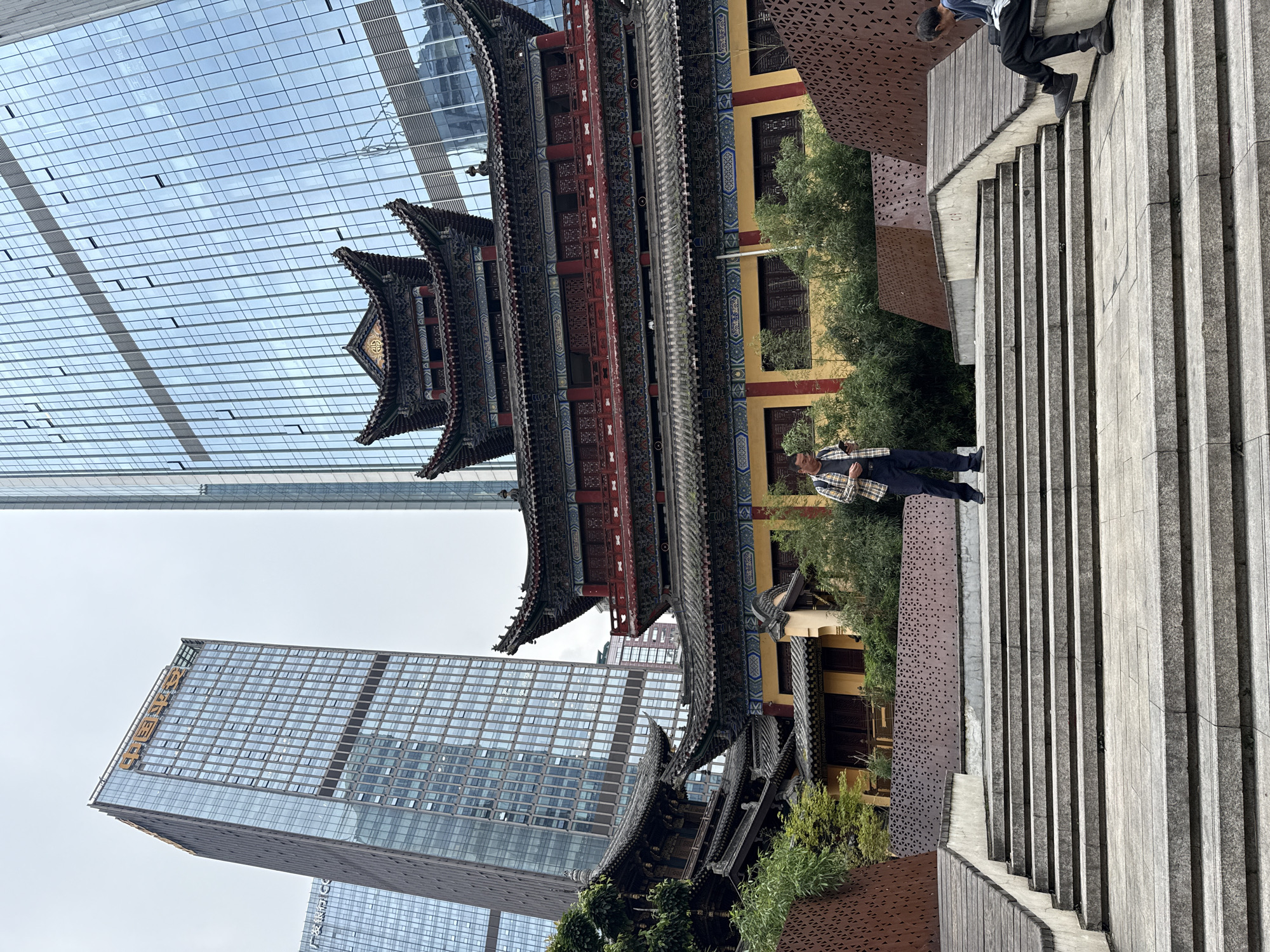 The image captures a juxtaposition of traditional Chinese architecture and modern skyscrapers in Yuzhong District, China. A multi-tiered pagoda-style building stands prominently in the foreground, showcasing intricate roof designs and ornate details. Adjacent to the pagoda, a modern glass skyscraper stretches towards the sky, reflecting the urban environment. A man stands on a set of stone stairs, dressed in a plaid shirt and dark pants, contributing a human element to the scene. The architectural details, the blending of old and new, and the presence of a person on the stairs provide a snapshot of Yuzhong District's urban landscape.