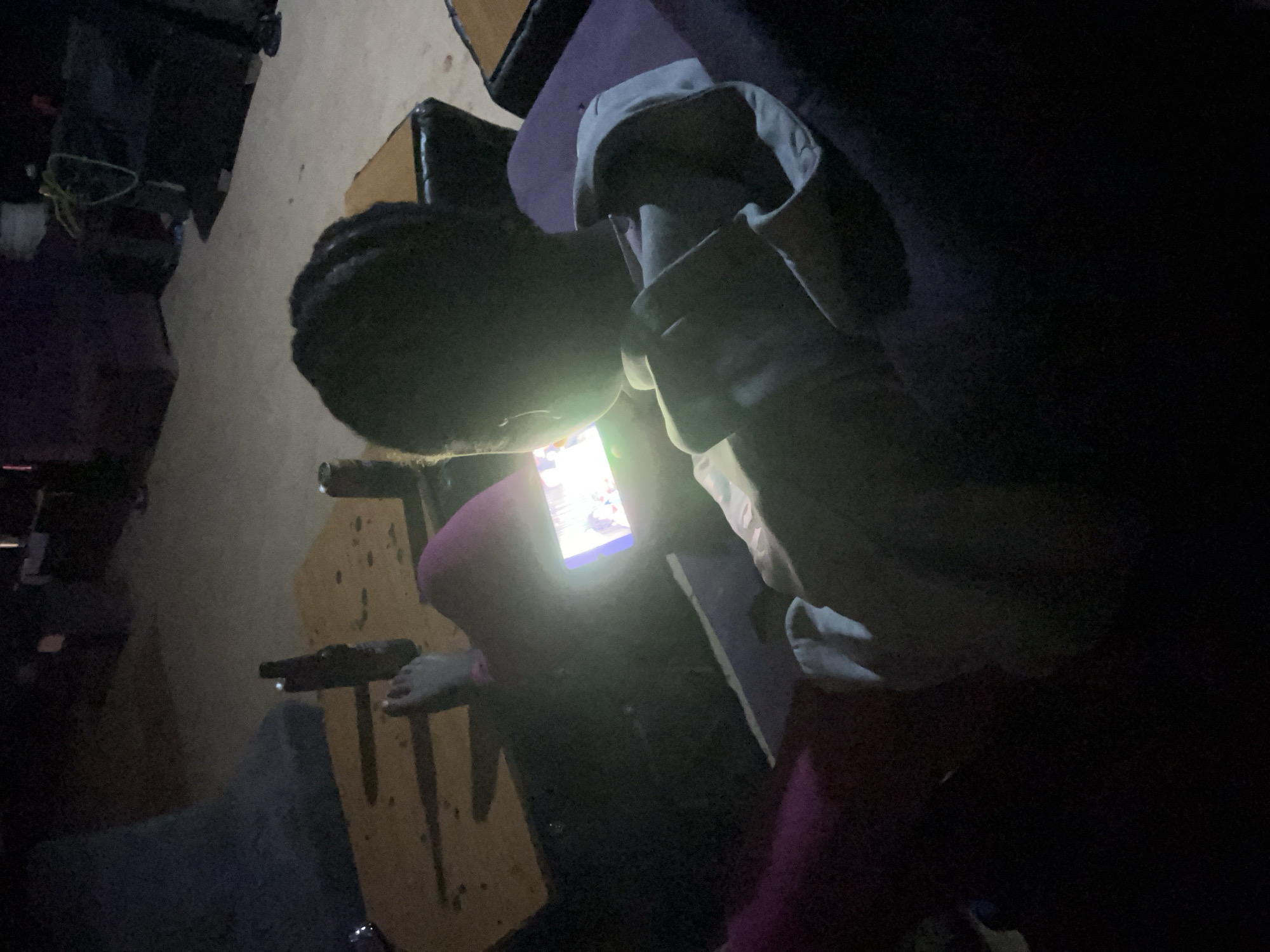 A dark, low-light image shows a person seated on a purple cushioned surface, looking at a smartphone. The person's head is partially obscured by darkness, but their braided hair is visible. They are wearing a light-colored shirt. Their legs are extended, and they are wearing dark pink leggings. The smartphone screen emits a bright light. To the left of the person, on a brown surface that appears to be a table, are two bottles, one dark and one lighter, and a can. The background is dark and indistinct, suggesting an indoor or dimly lit outdoor setting. The overall scene conveys a sense of quiet engagement with the digital device in a relaxed environment.