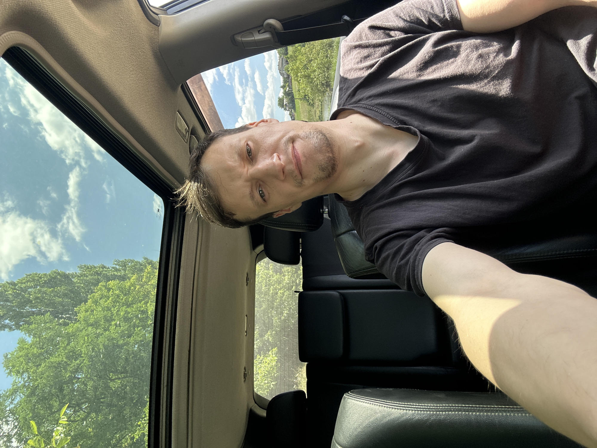 A man with short brown hair and stubble is sitting in the driver's seat of a car. He is wearing a dark t-shirt and is looking at the camera. The car has a sunroof open, revealing a bright blue sky with white clouds and green trees. The interior of the car is visible, including black leather seats and a portion of the dashboard. The background outside the car shows a rural landscape with houses and trees. The image appears to have been taken in Makiv, Ukraine, on a sunny day.