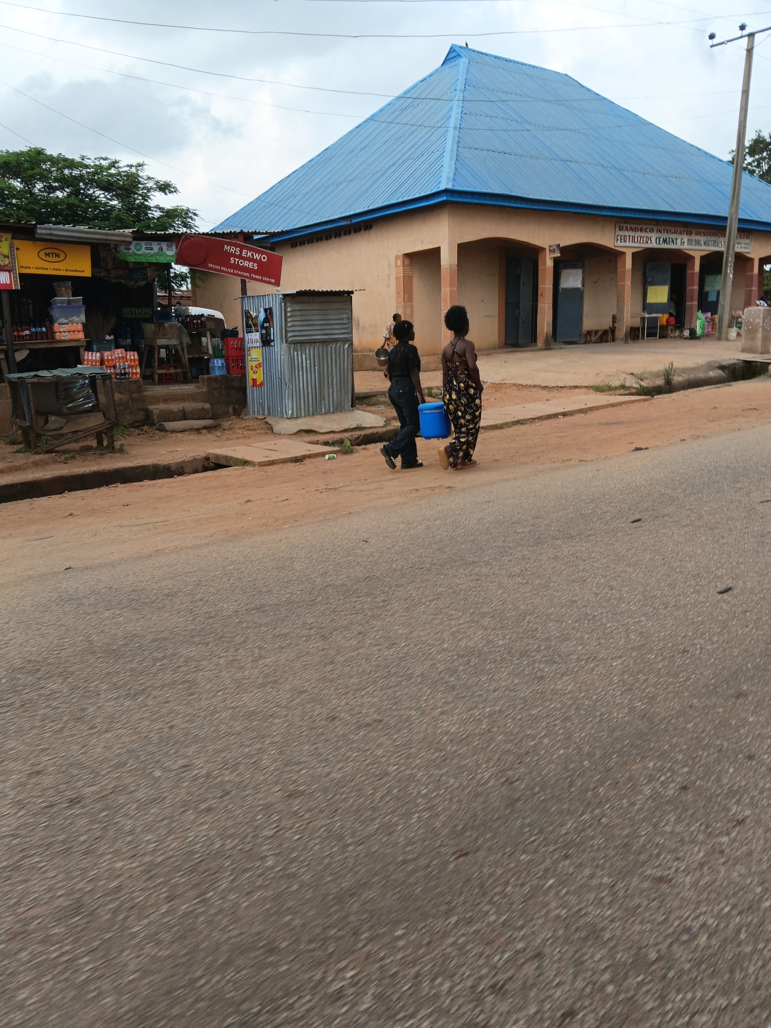 A medium shot captures a street scene in a Nigerian town, possibly in the afternoon given the lighting. The foreground is dominated by an asphalt road with a slight blur, suggesting the photo was taken from a moving vehicle. To the right, a dirt shoulder with some sparse vegetation leads to the background.
In the midground, a tan, single-story building with a bright blue, corrugated metal roof stands prominently. The building features several arched entrances. Above the entrances, a sign reads "BANDECO INTEGRATED RESOURCES LTD. FERTILIZERS, CEMENT & BUILDING MATERIALS". To the left of this building, a smaller shop with a red sign that says "MRS. EKWO STORES" is visible. This shop appears to be part of a complex with a corrugated metal structure. Goods are displayed on shelves outside this shop, including what appear to be bottles of drinks and other packaged items.
Two women are walking away from the camera on the dirt path in front of the buildings. The woman on the left wears dark clothing and jeans. The woman on the right is wearing a long, patterned dress and is carrying a large blue bucket. The overall environment suggests a commercial area in a developing urban or semi-urban setting. The sky is overcast with clouds.