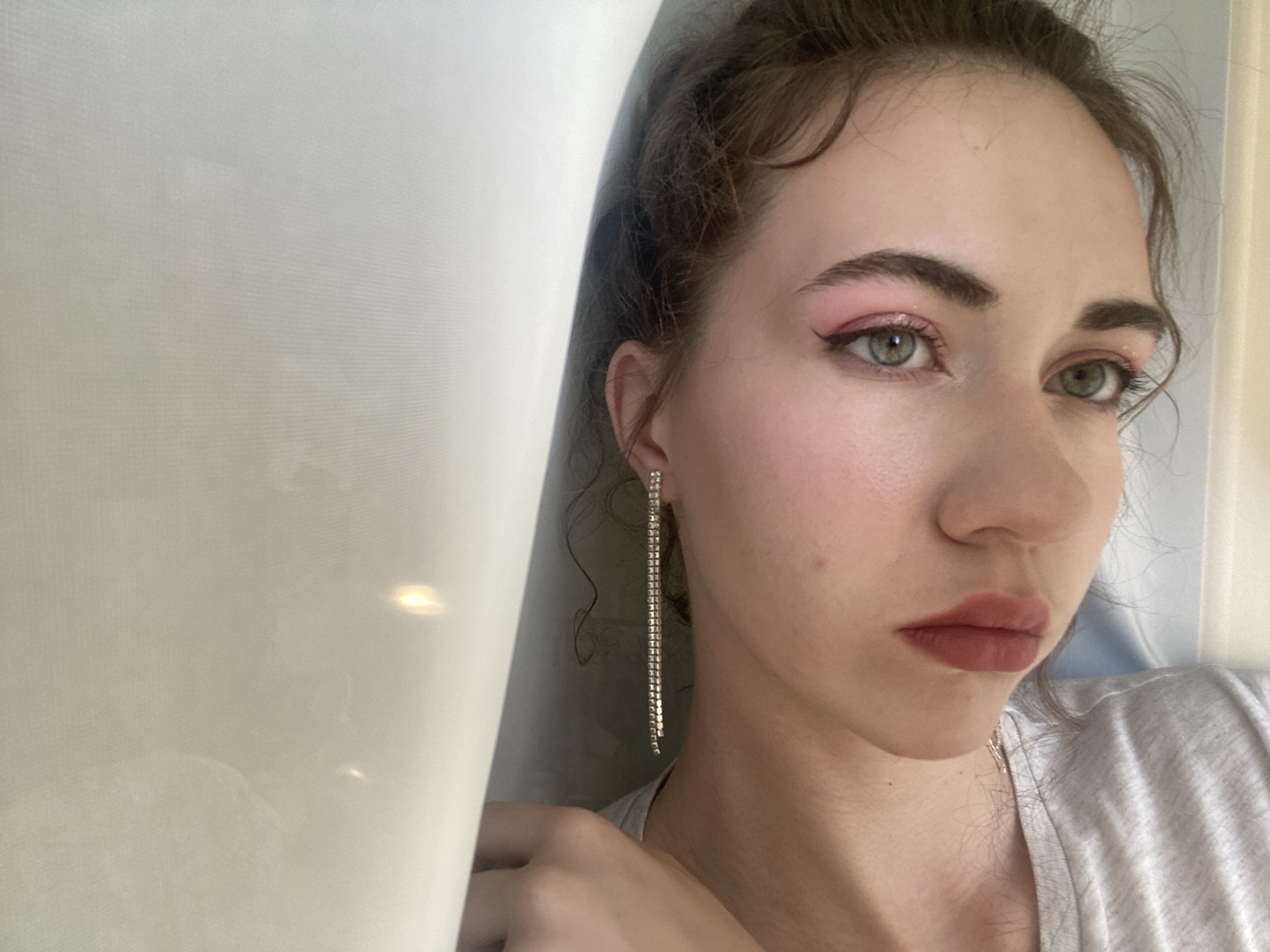 This is a medium shot, eye-level photograph of a young woman's face, cropped at the shoulders and positioned to the right of the frame. She is facing to the right, with her head slightly tilted down. Her gaze is directed downwards and away from the camera, giving her an introspective or perhaps slightly melancholic expression.
The woman has light skin, dark eyebrows, and dark hair pulled back in loose curls. Her eyes are a striking green. She is wearing makeup, with a noticeable pink eyeshadow that extends upwards towards her brow and a darker pink lipstick. A long, dangling silver earring with multiple rows of small, clear stones adorns her left ear. She is wearing a light grey t-shirt.
A large, pale, translucent curtain or fabric dominates the left side of the frame, obscuring about half of the image. The curtain appears to be made of a sheer material, possibly linen or a synthetic blend, with subtle horizontal lines. Light is filtering through the curtain, creating a soft, diffused illumination on the left side of the image, while the right side of the woman's face is more directly lit, revealing some subtle skin imperfections and the texture of her makeup.
The overall mood is subdued and intimate, with the soft lighting and the woman's downcast gaze contributing to a sense of quiet contemplation. The background is otherwise nondescript, with the curtain and a hint of a lighter wall or doorframe to the right. There is no visible text in the image. The implied location context is Zavodoukovsk, Russia, though this information is not visually evident in the photograph itself.