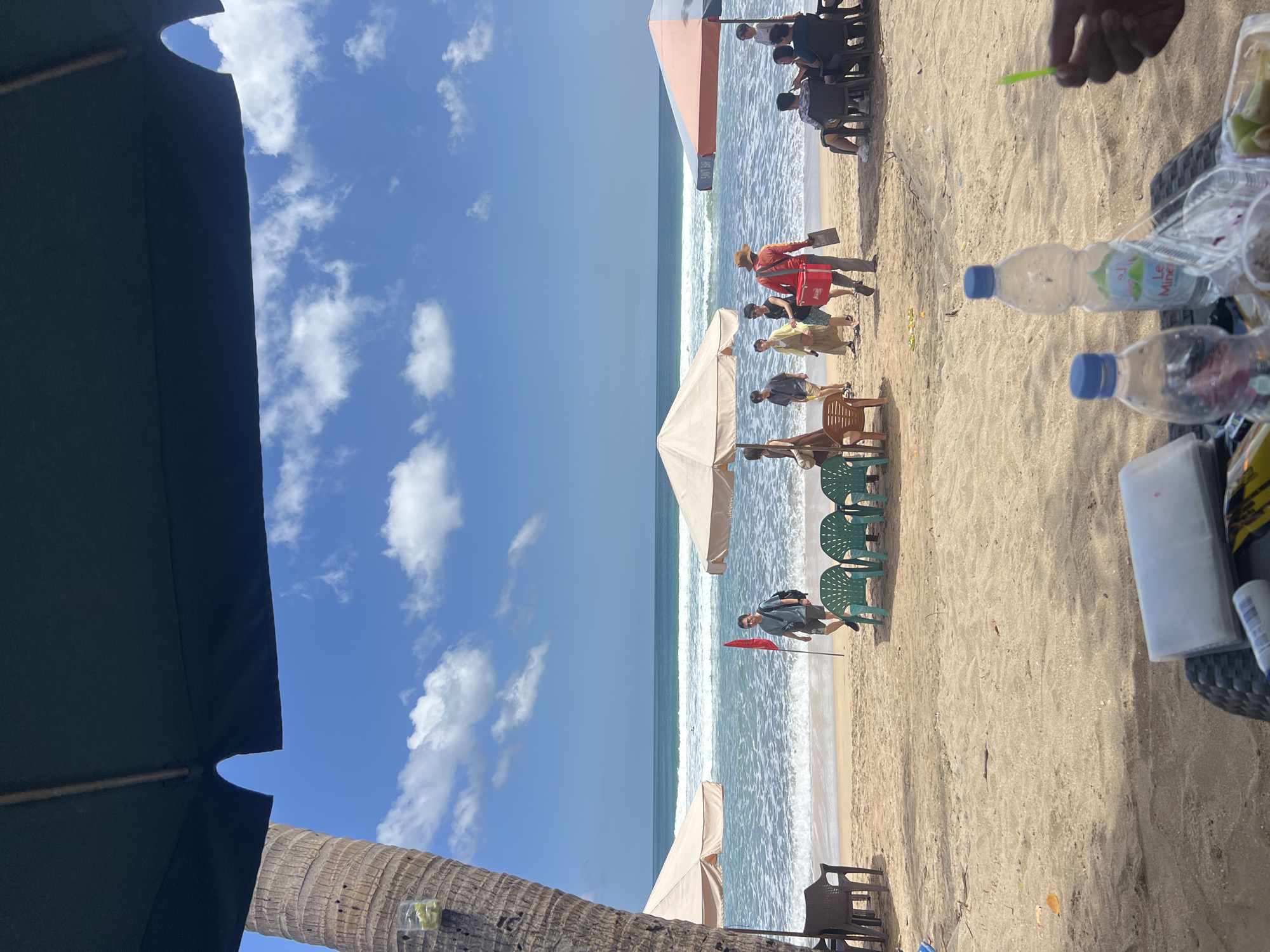 A group of people are enjoying a day at Kuta Beach, Indonesia, with the ocean and blue sky in the background. In the foreground, a person's hand is visible holding a green object, possibly a lime wedge. Two plastic water bottles and other items are on a table. Beach chairs and umbrellas are set up on the sand. Several individuals are walking along the beach, some are gathered under umbrellas, and others are seated. A man in a red shirt and hat is walking with a red cooler bag. The waves are gently breaking on the shore.