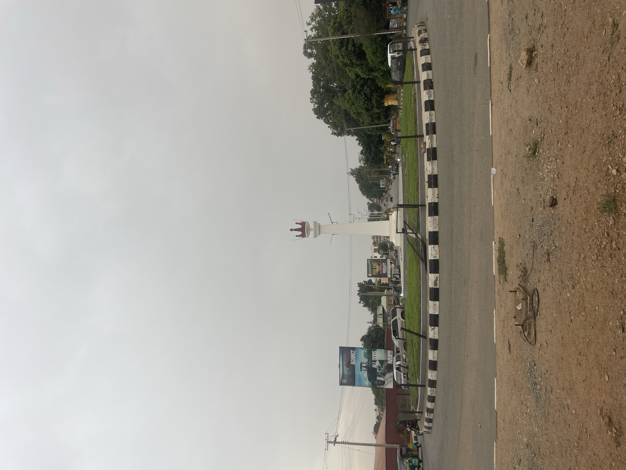 The image depicts a street scene in Jos, Nigeria, featuring a prominent white tower topped with a red crown-like structure, presumably a landmark. The sky is overcast. Along the right side of the frame is a paved road with a black and white striped curb. Beyond the curb, a dirt area stretches to the edge of the frame, where a broken bicycle frame is visible. Adjacent to the road is a grassy median with sparse vegetation. In the background, buildings, trees, billboards, and vehicles are seen, indicating urban activity. A large billboard displays an advertisement. A tricycle is on the road.