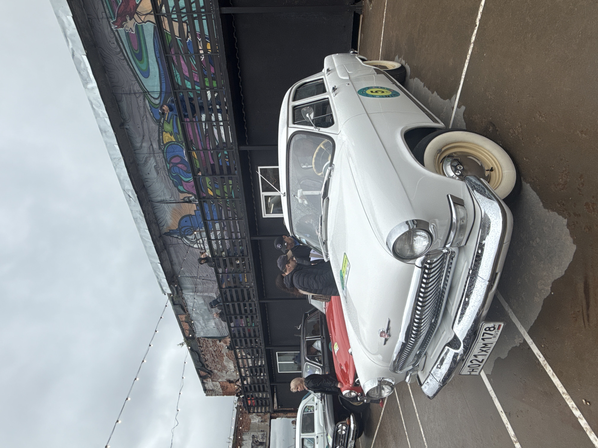 The image shows a row of vintage cars lined up on a wet street in Saint Petersburg, Russia. The cars are of different colors, including white, red, and black. The white car in the foreground is a GAZ-21 Volga, with a Russian license plate "H021XM 178". Several people are standing around the cars, possibly admiring them or taking pictures. A building with a mural on its side can be seen in the background, along with exposed brick and string lights. The sky is overcast, suggesting it might be raining.