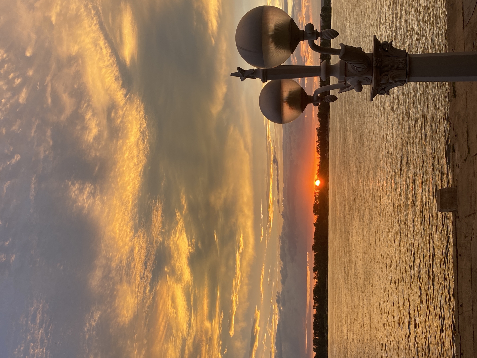 A sunset over the Volga River in Novokuybyshevsk, Russia, is captured in this vertical, eye-level, medium shot. The sky is ablaze with vibrant oranges and yellows, with streaks of grey clouds adding contrast. The sun, a bright orange orb, is partially visible on the horizon, casting a warm glow across the water. A dark, tree-lined shoreline stretches across the middle ground. In the foreground, a lamppost with two globe-shaped lights stands to the right, its ornate design visible. The water's surface is rippled, reflecting the colors of the sky. A concrete embankment with a small, block-like object is in the lower foreground. There are no people or animals visible in the scene.