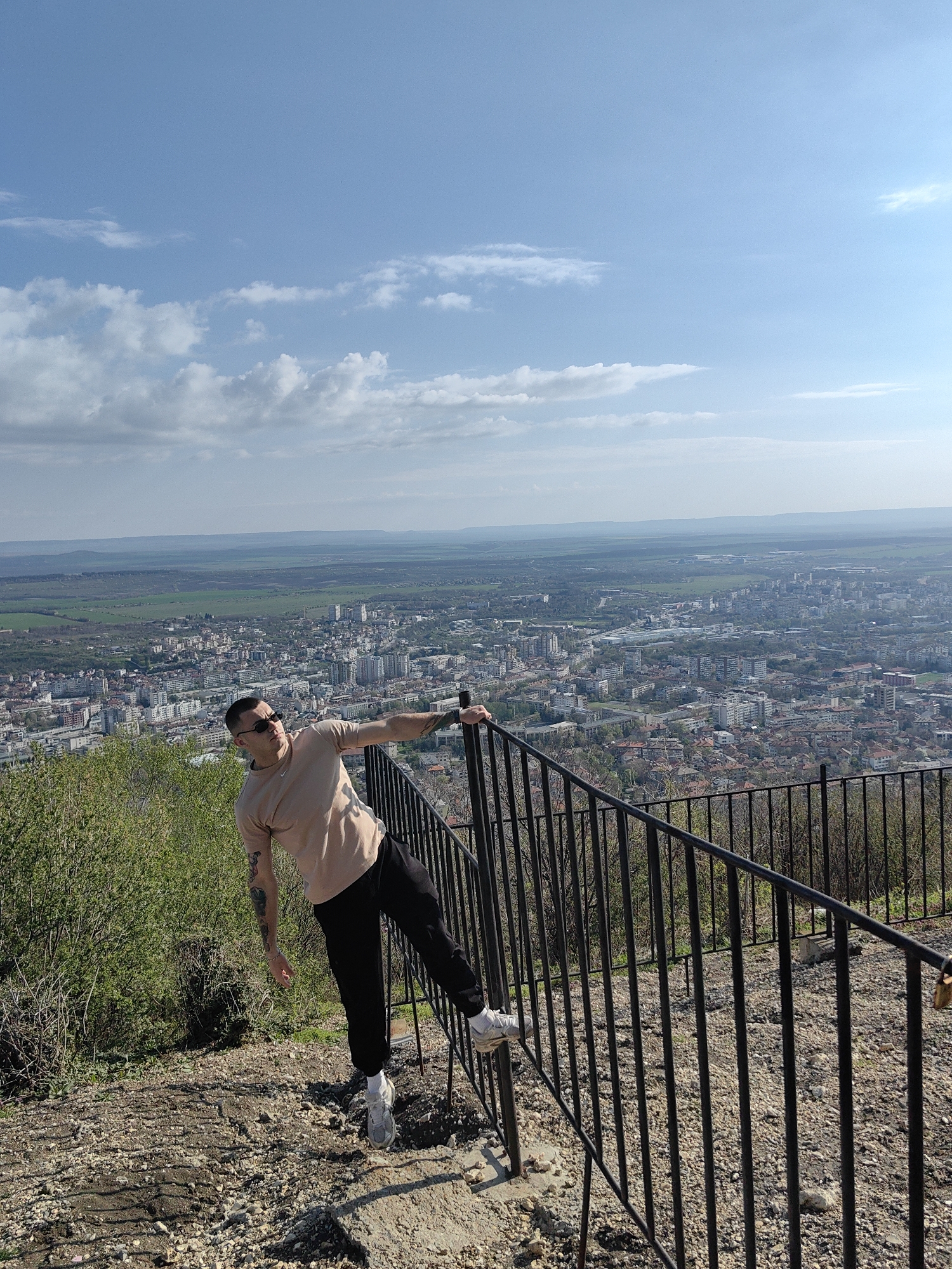 A man is standing on a hill overlooking Shumen, Bulgaria. He is wearing sunglasses, a light brown t-shirt, black pants, and gray shoes with white socks. He is leaning on a black iron fence. The fence is constructed of vertical bars. To the left of the man is a green bush. The foreground is a rocky, dirt area. In the distance is the city of Shumen, with buildings and trees visible. The sky is blue with white clouds.