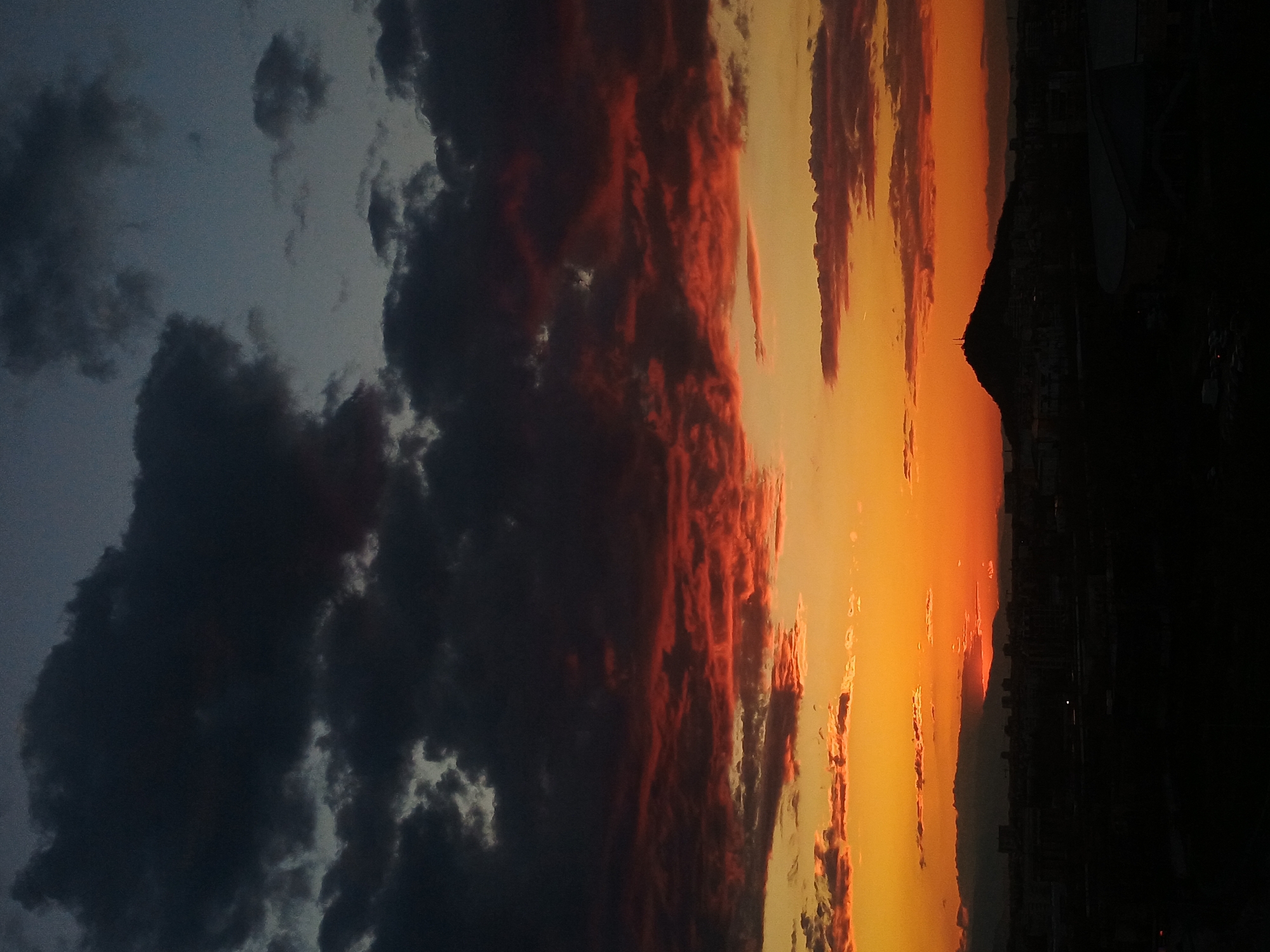 The image shows a sunset in Plovdiv, Bulgaria. A mountain silhouette is visible on the right side of the frame, contrasting against a bright orange and yellow sky. A city skyline is silhouetted at the base of the mountain. To the left, a mix of dark grey and red-tinged clouds fills the sky. Some buildings are visible on the right of the frame. The photograph is oriented sideways, with the sky on the right and the mountain on the bottom.