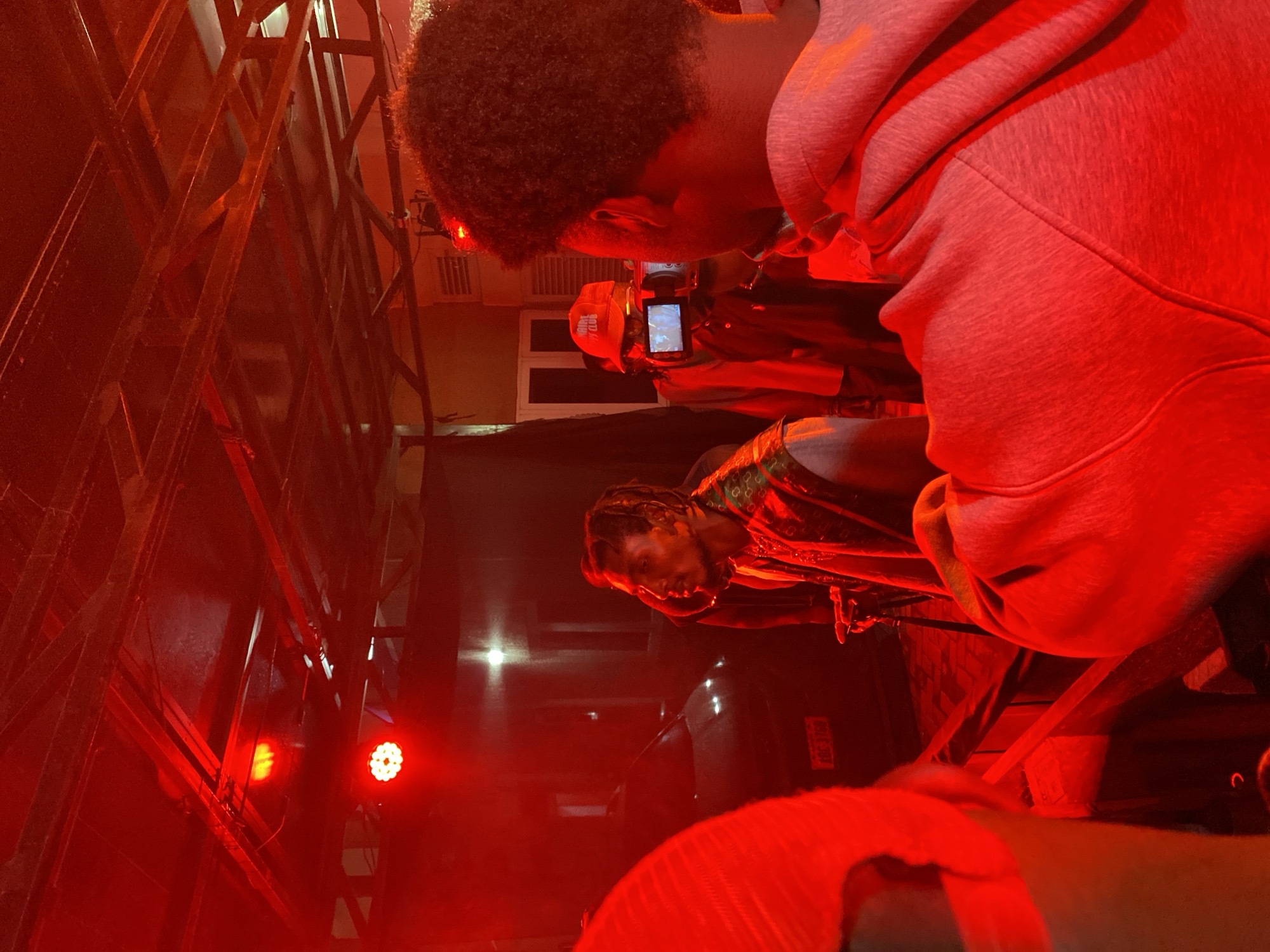 In Abuja, Nigeria, a group of individuals are gathered in a dimly lit environment, dominated by red lighting, likely a studio or event space. The scene is focused on a man with dreadlocks, wearing a patterned vest over a white shirt, who is captured in profile, looking to his right. He appears to be in conversation or interacting with someone outside the frame. To his right, another individual, seen from the back, holds a video camera, potentially recording the man. This person wears a red baseball cap with the logo "RIDE CLUB" visible. In the foreground, a person is partially visible on the left, their red attire creating a vibrant contrast, and another person in the foreground on the right, seen from the back, wears a grey hooded sweatshirt. In the background, a dark-colored car is visible, suggesting an outdoor or semi-outdoor setting, possibly a driveway or parking area. The overall atmosphere is that of a filming or recording session.