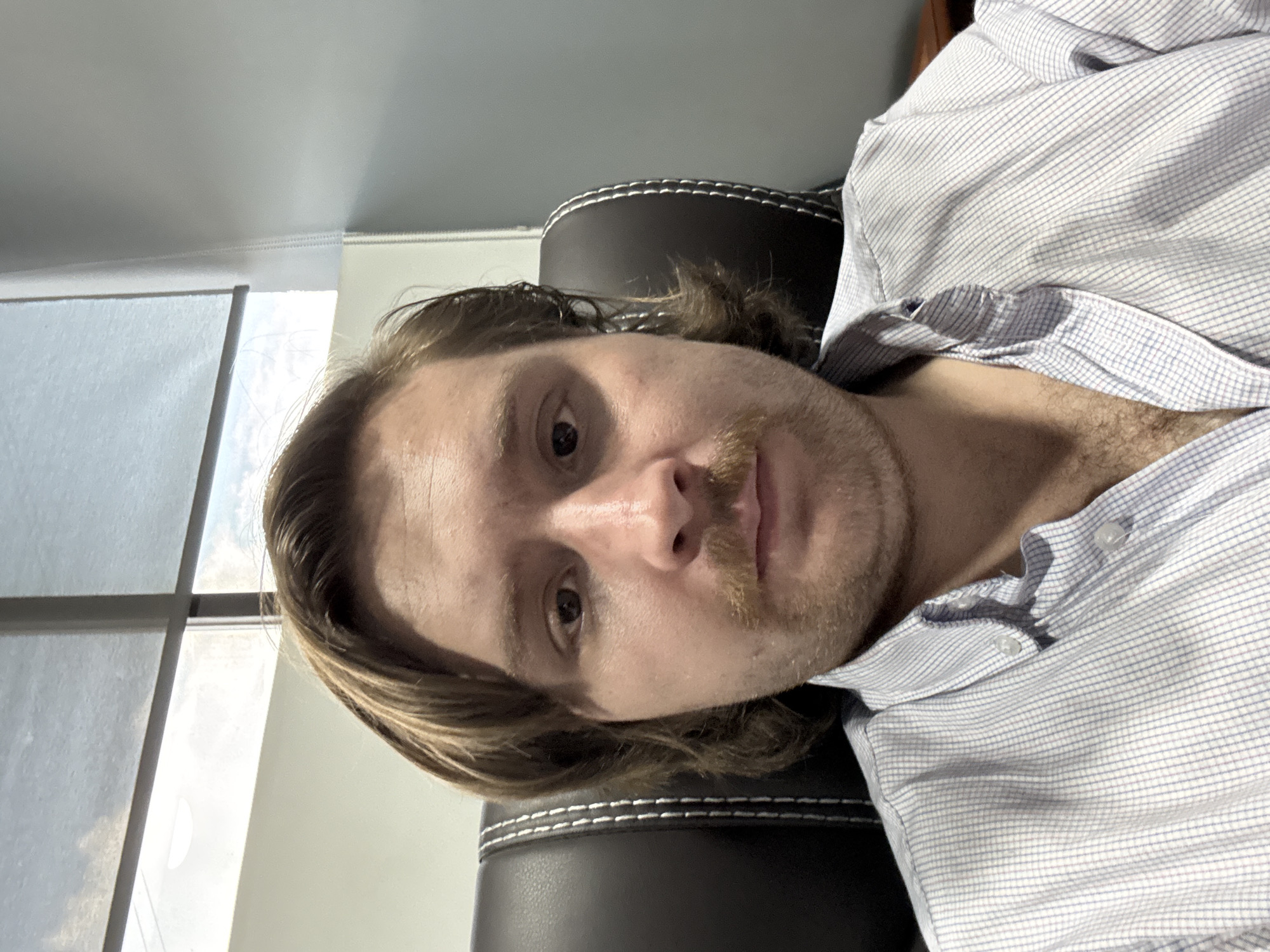 A person is seen from a close, downward angle. The person has light brown hair, a mustache, and appears to be wearing a blue and white patterned button-down shirt. The person is sitting in what seems to be a black chair with white stitching. A window is to the left of the person with reflected light. The location is not indicated in the photograph.