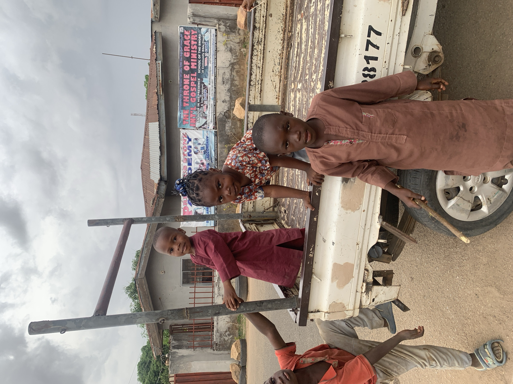 The image shows three children and a man on or around the bed of a white truck with the partial license plate "8177". The background contains buildings and signage in the city of Jos, Nigeria. Specifically, there are posters for "The Throne of Grace INT'L Gospel Ministry" and "Rose Stone Academy". The children appear to be playing on or near the back of the truck, while the man is partially visible beneath the truck bed. The sky is overcast.