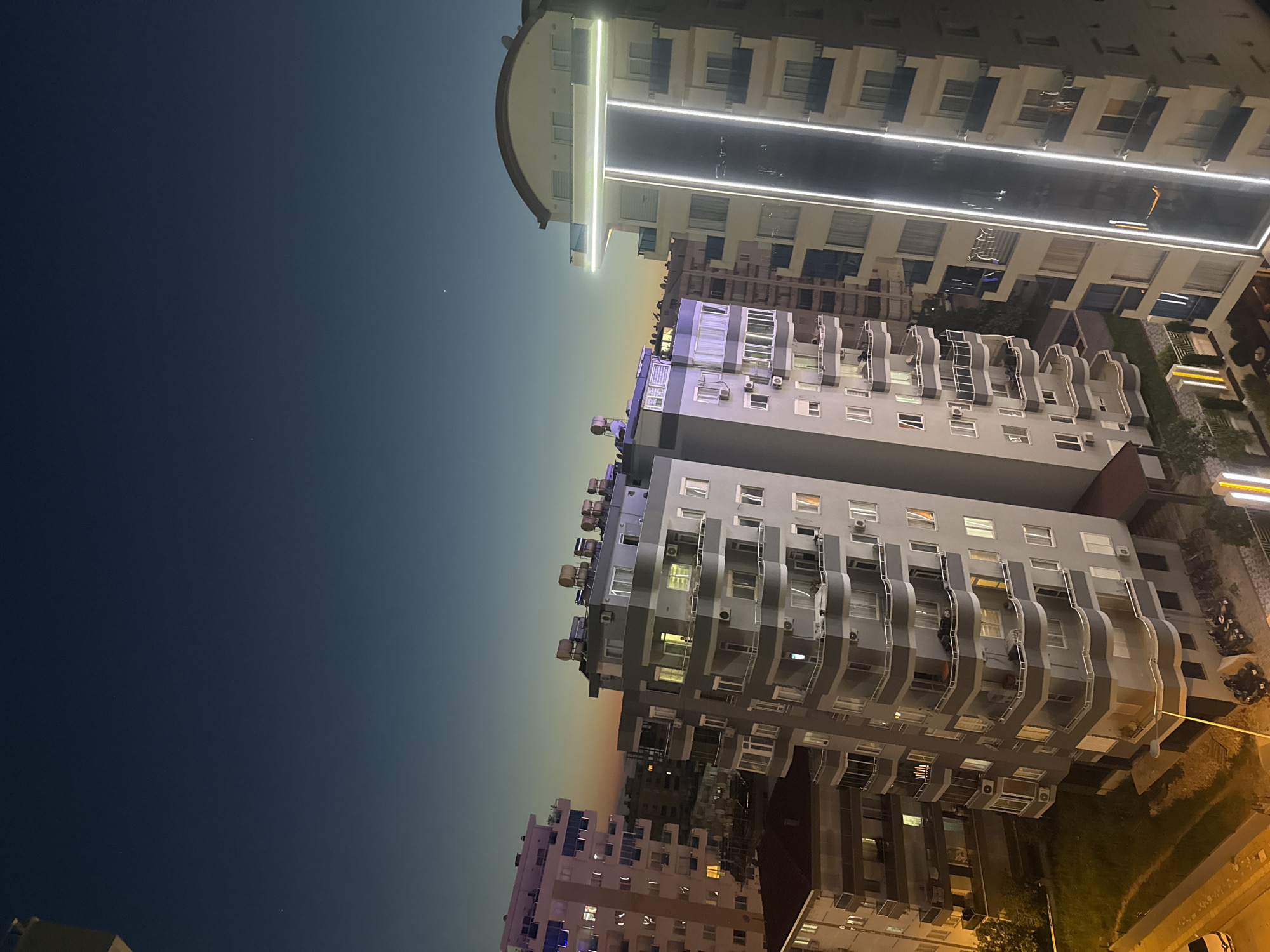 The image displays an urban landscape captured at night or during late twilight from an elevated viewpoint. The sky gradients from a deep, dark blue-grey at the top to a softer, yellowish-orange hue near the horizon, suggesting either the last vestiges of sunset or significant light pollution. A few faint, bright specks, possibly stars or distant lights, are scattered across the darker sky.
Multiple multi-story buildings are the primary subjects. In the upper left area of the correctly oriented image, a prominent, modern building with a rounded roof stands out, featuring several horizontal strips of bright white LED lights illuminating its facade. Below and to the right, a cluster of other buildings, mainly grey or light grey, is visible. These structures are characterized by numerous balconies, many exhibiting curved or wave-like designs, and an abundance of windows. Some windows glow with warm interior lights, while others remain dark. Exterior air conditioning units are visible on many walls and balconies.
At the lower right, the ground level shows patches of green grass, possibly a paved area, and several parked vehicles, including what appear to be motorcycles and a white car. This area is lit by bright, yellowish artificial lights.
No people or discernible activities are visible in the scene. There is no legible text present on any of the buildings or objects. The overall impression is a quiet, illuminated cityscape after dusk.
