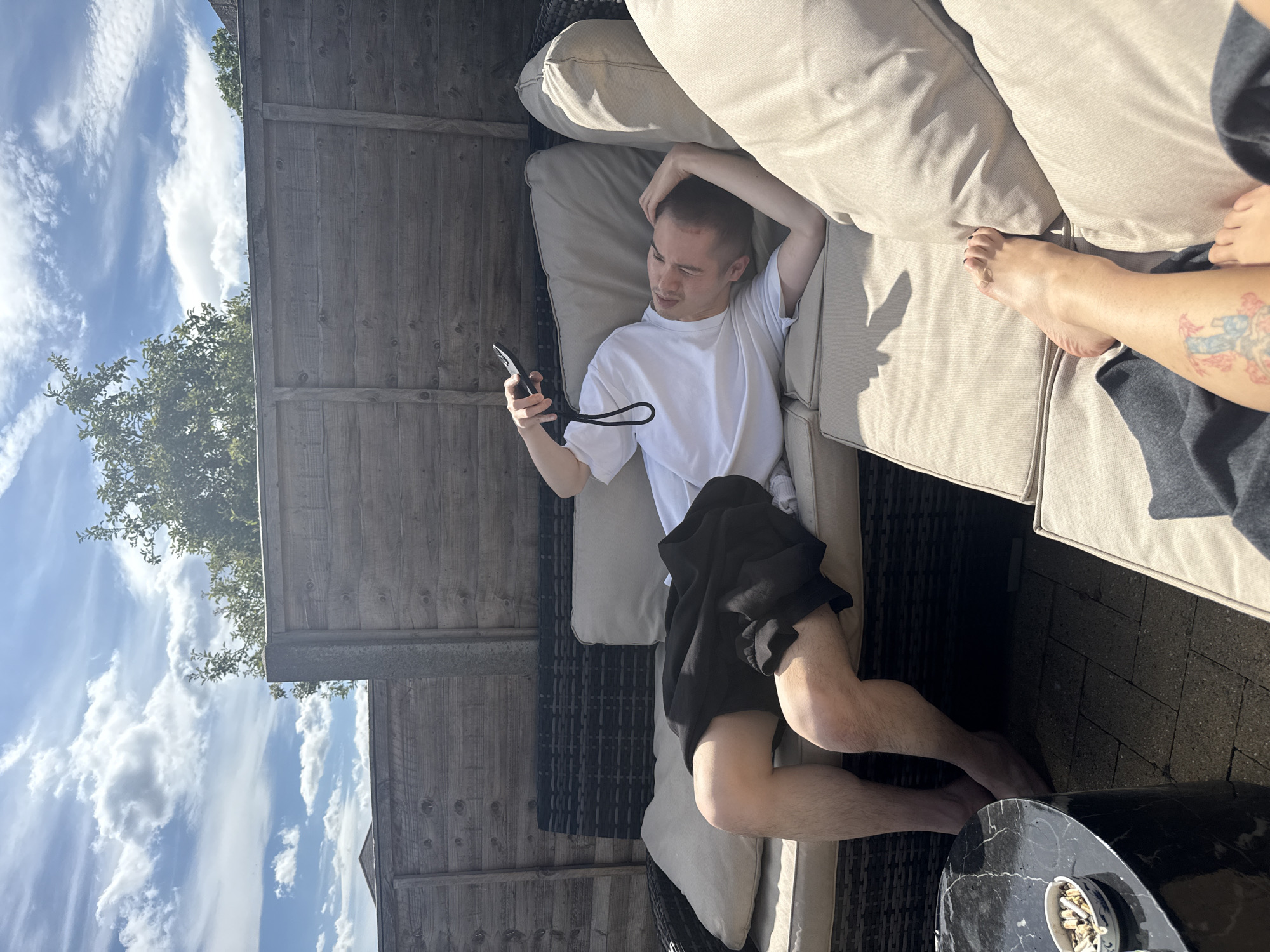 A young male is lying on a cushioned outdoor sofa, holding a remote control and looking up at the sky. He is wearing a white t-shirt and black shorts. The sofa is situated on a paved patio area, next to a wooden fence. In the foreground, a black marble coffee table is visible, with an ashtray filled with cigarette butts on its surface. The sky is cloudy with patches of blue, and trees are visible behind the fence. The scene appears to be a residential garden.