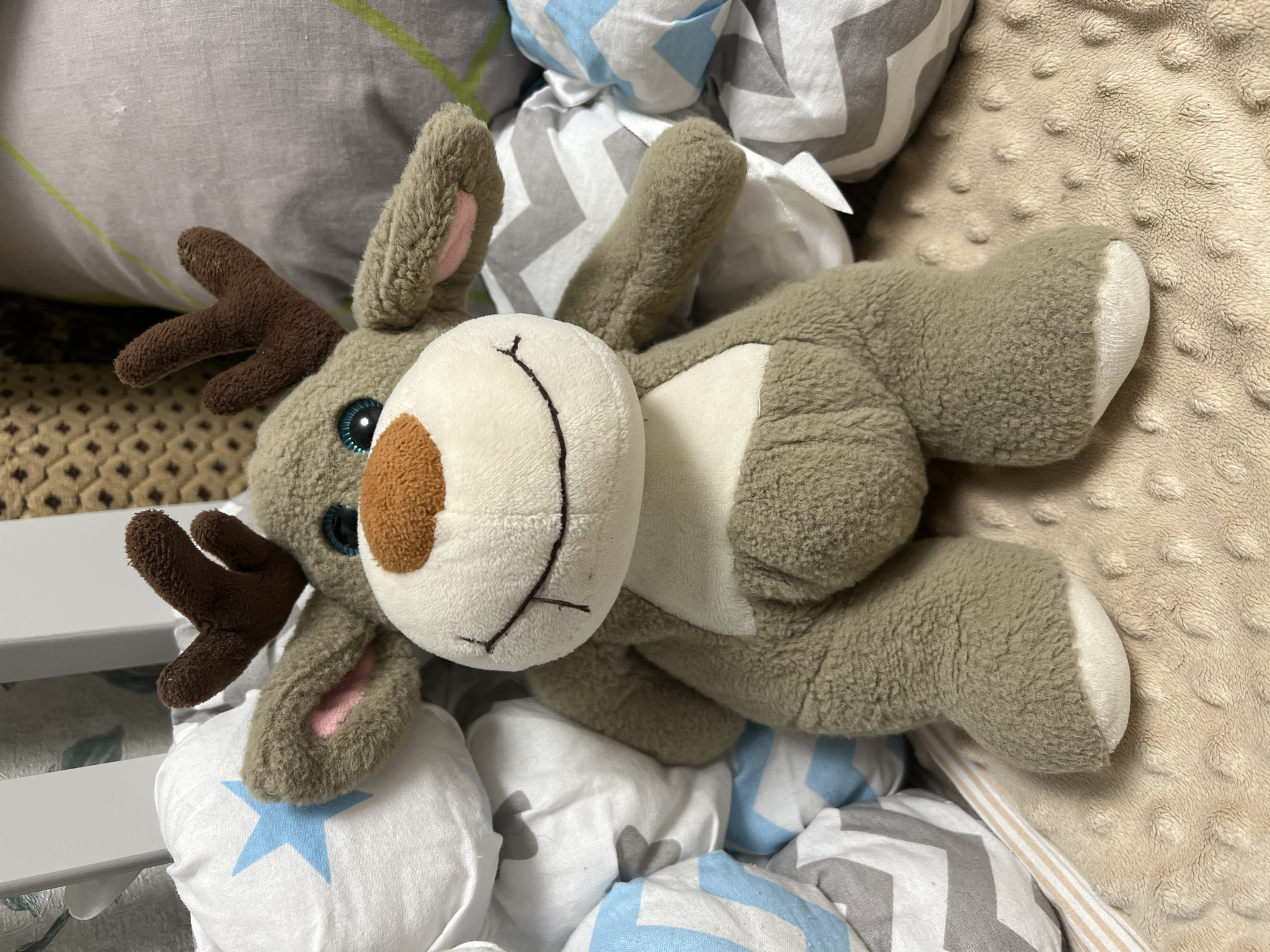 A stuffed reindeer toy with brown antlers and blue eyes is sitting amongst patterned pillows and a white crib railing. The reindeer is facing forward and has a white belly and a brown nose. The pillows are white with blue and gray zig-zag patterns, and some have a blue star. The environment appears to be a nursery or child's bedroom. The location is Unknown, Unknown.