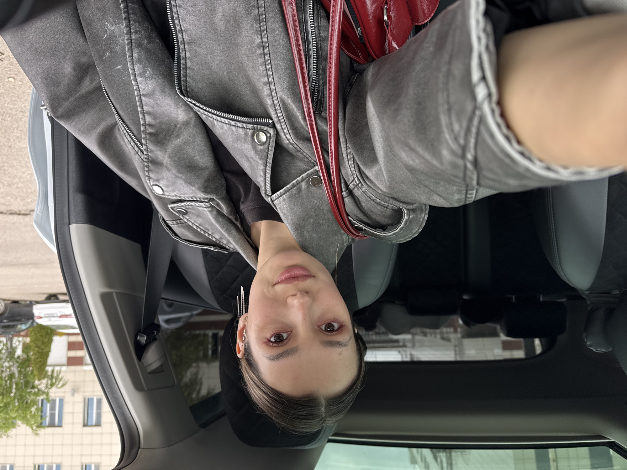 A woman is seated in the driver's seat of a car. She is wearing a gray leather jacket and a red purse is hanging on her shoulder. The interior of the car is visible, including the seats and dashboard. Outside the car, there is a building and a tree.