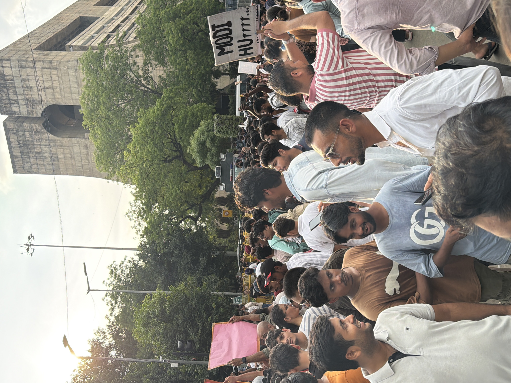 The image depicts a crowd of people gathered on a street in New Delhi, India, with a large, Brutalist-style building in the background. Many individuals are holding signs and appear to be participating in a protest or demonstration. The atmosphere seems energetic, with people looking in various directions and engaging with their surroundings. The sun is casting a warm glow, suggesting it is either morning or late afternoon. Streetlights and trees are visible, contributing to the urban environment.