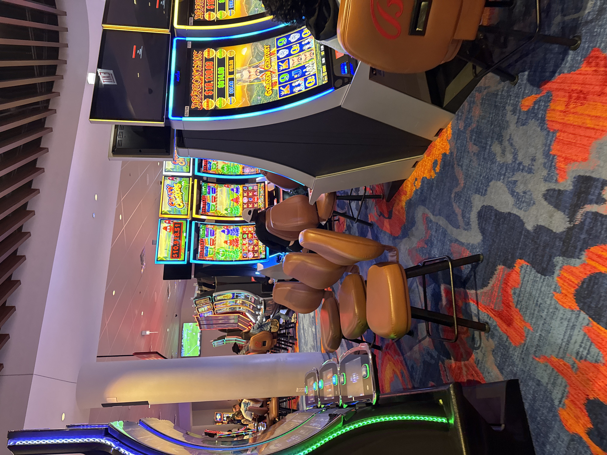 The image shows an interior view of a casino. Rows of slot machines are visible, illuminated with bright displays. Some machines feature games like "CRAZY CHICKENS" and "DRAGON LINK," displaying jackpot amounts. The flooring is a patterned carpet in shades of blue, orange, and gray. Several brown cushioned chairs are positioned in front of the machines. People are seen seated at some of the slot machines. The environment appears to be well-lit with modern architectural details like a curved counter with green LED lighting. The location can not be determined to be in the city of Chicago, United States of America with the given context.