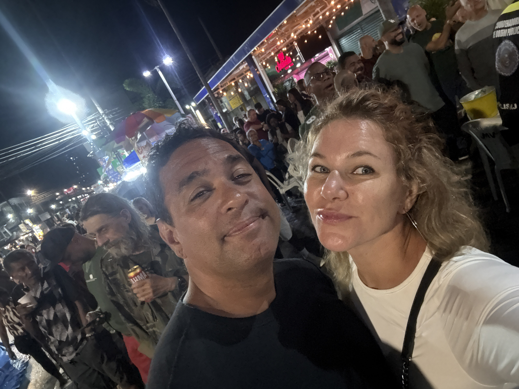 The image is a selfie taken at night, possibly at a street festival or outdoor event. Two people, a man and a woman, are prominently featured in the foreground, smiling at the camera. The man is wearing a black t-shirt, and the woman is wearing a white long-sleeved shirt.
In the background, a crowd of people are visible. They appear to be enjoying themselves, with some interacting with each other. There are street vendors with umbrellas and lights, suggesting a festive atmosphere. The environment appears to be an urban setting with buildings and streetlights.
While the image itself doesn't explicitly state the location as Nilópolis, Brazil, it aligns with descriptions of street events that occur in cities like Nilópolis, which are known for their vibrant nightlife and community gatherings. The presence of diverse individuals and the lively atmosphere are characteristic of such events.