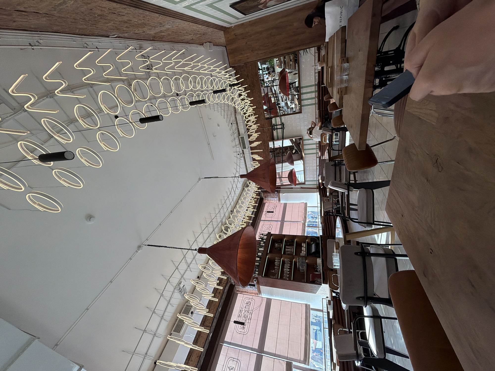 The image shows the interior of a restaurant with a modern design, located in Saint Petersburg, Russia. The ceiling features an intricate array of neon ring lights and zig-zag patterns, as well as cylindrical pendant lights. Large windows with beige blinds line the left side of the room, letting in natural light. A tall wooden shelving unit filled with bottles and glassware stands near the window.
The restaurant is furnished with various wooden tables and chairs. In the foreground, a person's hands are visible, holding a dark rectangular object, possibly a phone. In the background, several patrons are seated at tables, some appear to be dining, while one person is looking at a document or menu. The walls are a mix of exposed brick and tiled sections with green and white geometric patterns. The overall atmosphere suggests a contemporary dining establishment.