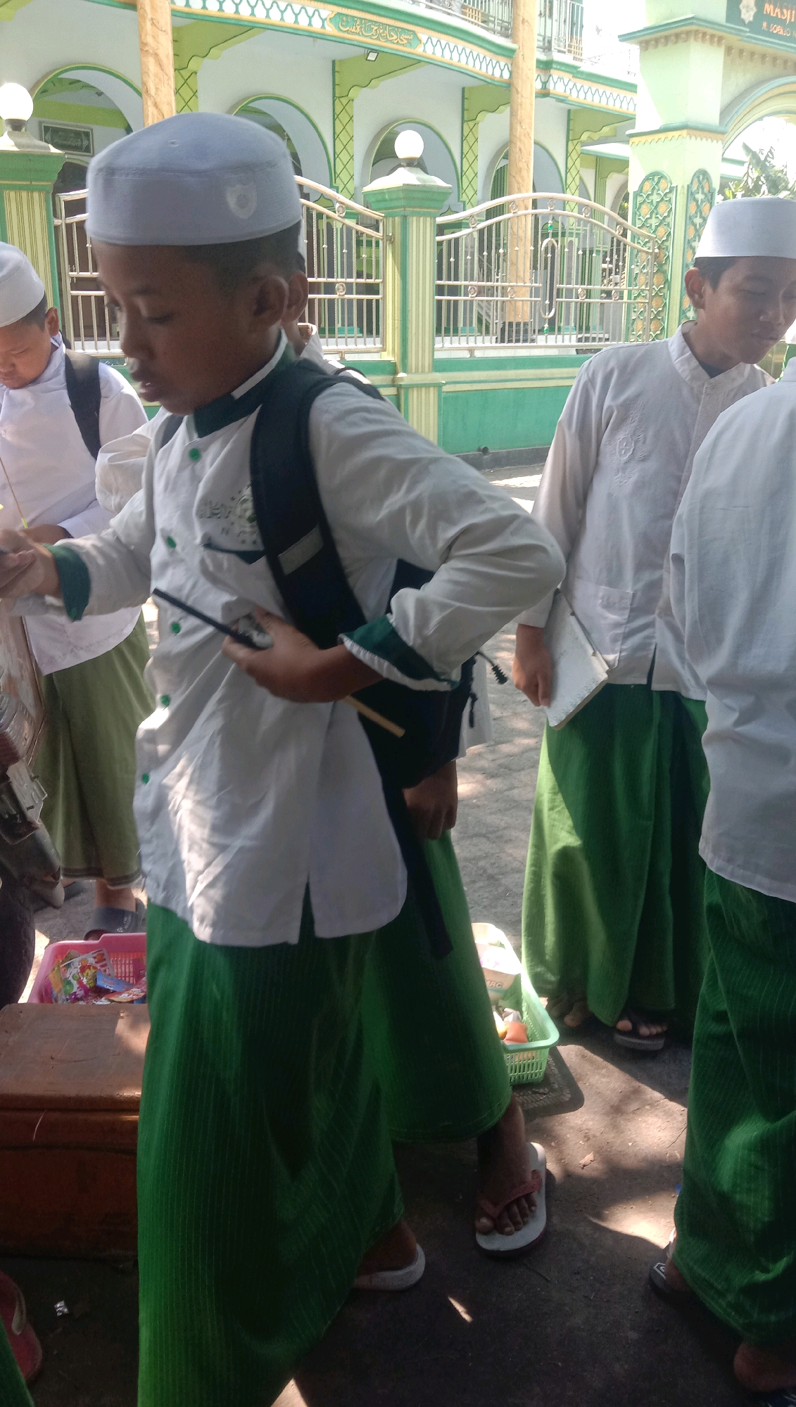 A group of young boys, likely students, are gathered outside a mosque in Ngempit, Indonesia. They are dressed in traditional Islamic attire, consisting of white long-sleeved shirts, white skullcaps, and green sarongs or trousers. The mosque, with its green and white architectural details and arched entrances, serves as the backdrop. One boy in the foreground is holding a thin stick and appears to be engaged in an activity or conversation. Another boy next to him is also looking down, possibly interacting with him. The ground is paved with concrete, and sunlight casts shadows, indicating it is daytime. A wooden crate and a green plastic basket containing various items are visible on the ground. The overall scene suggests a cultural or educational gathering related to the mosque.