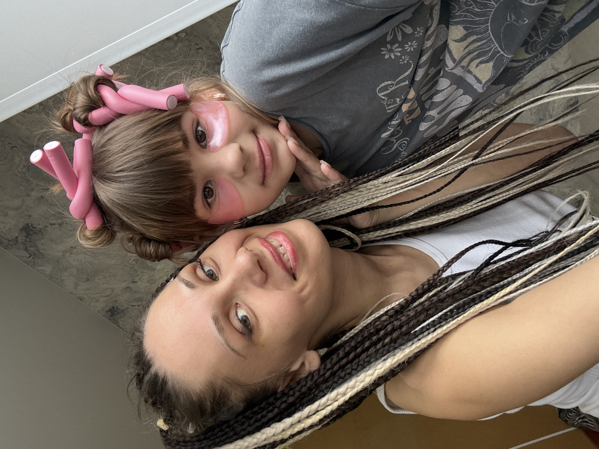A close-up, high-angle selfie features two females, an adult and a child, lying down indoors and looking at the camera. The younger individual, positioned above the adult, has light brown hair with bangs, styled with numerous pink foam hair rollers. She wears pink under-eye patches and a gray t-shirt with a decorative pattern featuring a sun, flowers, and abstract shapes. She displays a soft smile.
Below her, the adult smiles warmly. Her hair consists of long, intricate braids in varying shades of dark brown, black, and light blonde or white. She is wearing a white ribbed top. Both individuals appear relaxed and content.
The scene suggests a casual, personal moment, possibly a shared beauty or pampering routine, given the child's hair accessories and eye patches. The background is simple and out of focus, indicating an indoor environment with plain surfaces. No specific location, time of day, or weather can be discerned. There is no visible text in the image.