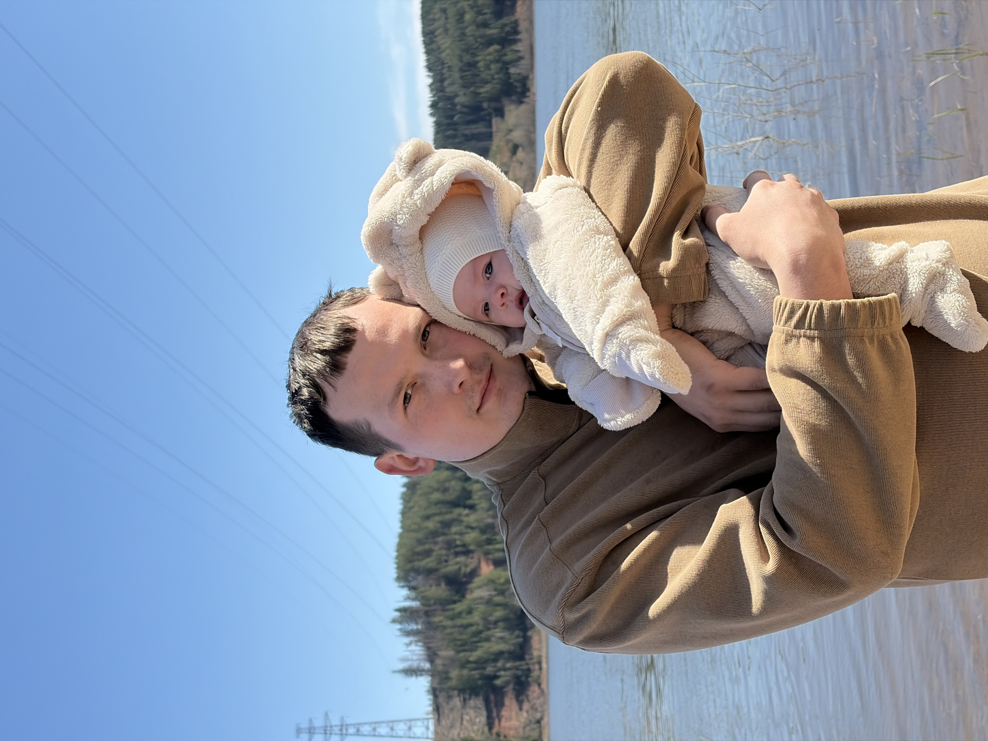 A medium shot depicts a man holding a baby outdoors near a body of water. The man is wearing a brown, long-sleeved shirt. The baby is clad in a white, fuzzy, hooded outfit. Power lines are visible in the sky, with a support tower partially seen. A forested hillside rises in the background. The water suggests a lake or large river, with reflections distorting the image. The location is the city of Chaikovsky, Russia.