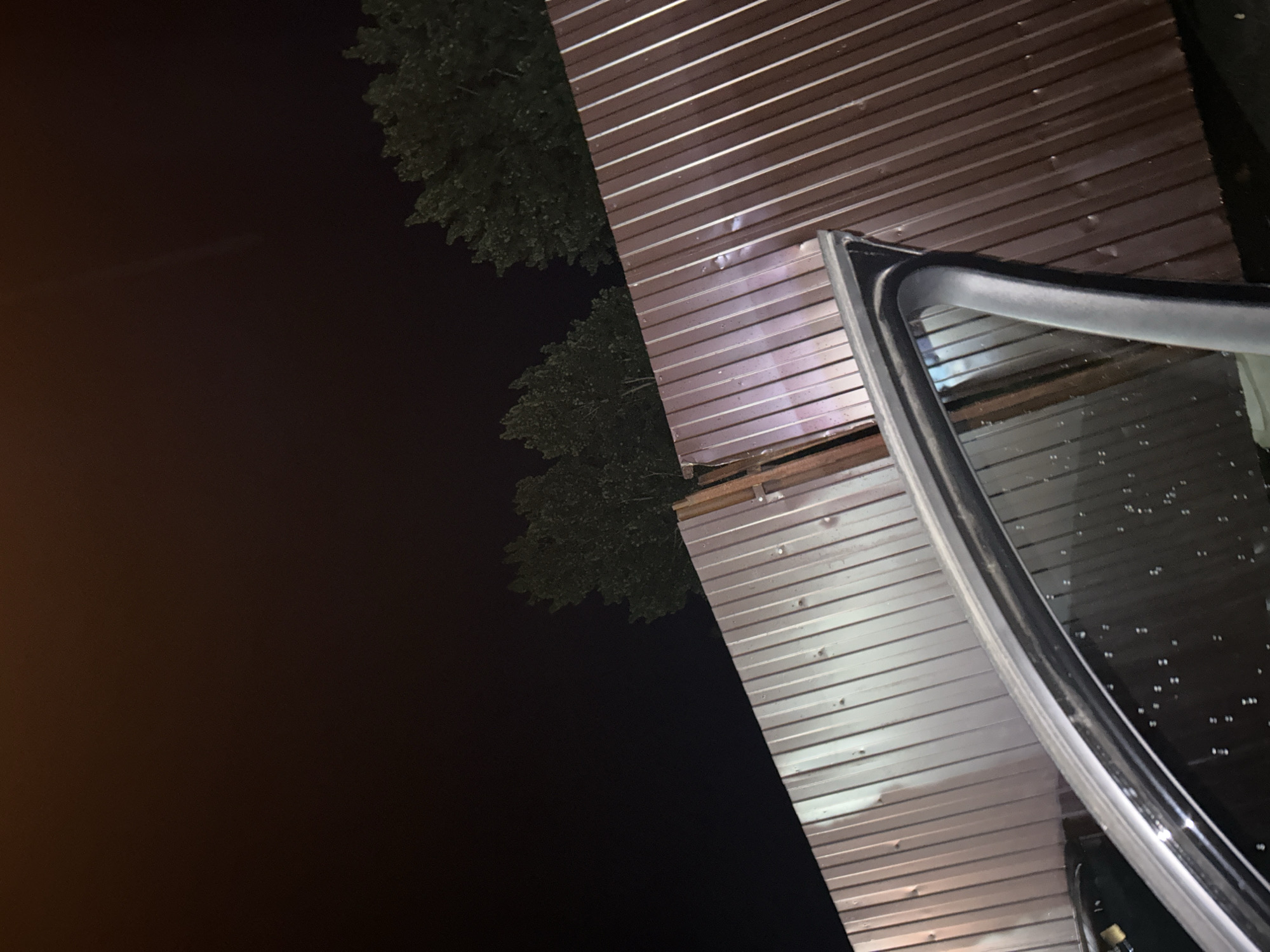 In a dark, urban environment, a corrugated metal fence is visible. The fence is primarily brown and appears damaged in the center, with a section bent and distorted. A car door is open and positioned in the foreground, partially obscuring the fence. The sky above is black, suggesting nighttime. Trees are visible in the background, silhouetted against the dark sky. The scene is devoid of human presence.