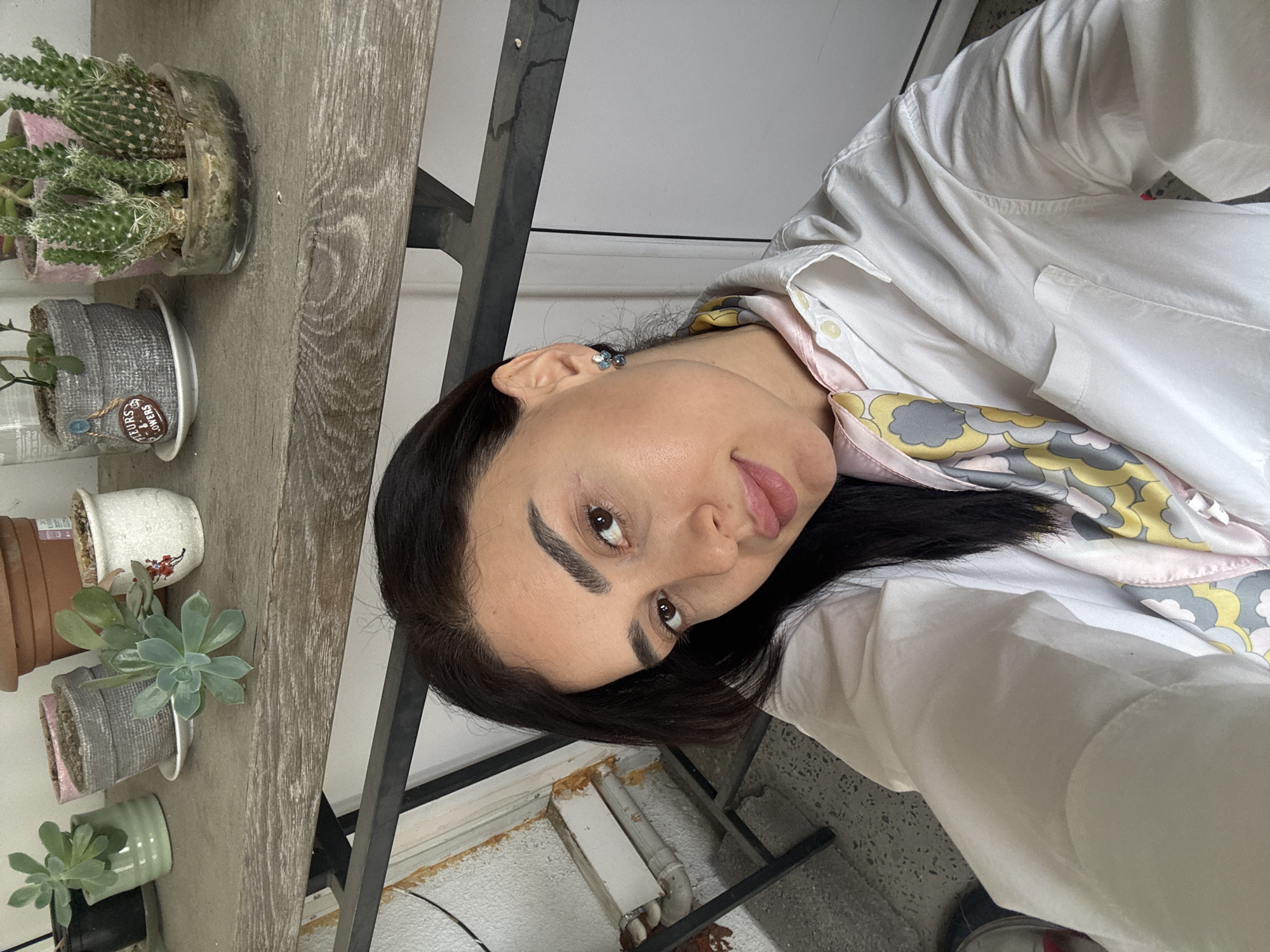 A selfie shows a woman wearing a white shirt and a patterned scarf, posed near a wooden shelf holding several potted plants, including cacti and succulents. The location is Tbilisi, Georgia, suggested by architectural features in the background and possibly the style of the building. The woman is indoors, in a space with a balcony or terrace as hinted by the open window in the background. The lighting appears to be natural daylight.