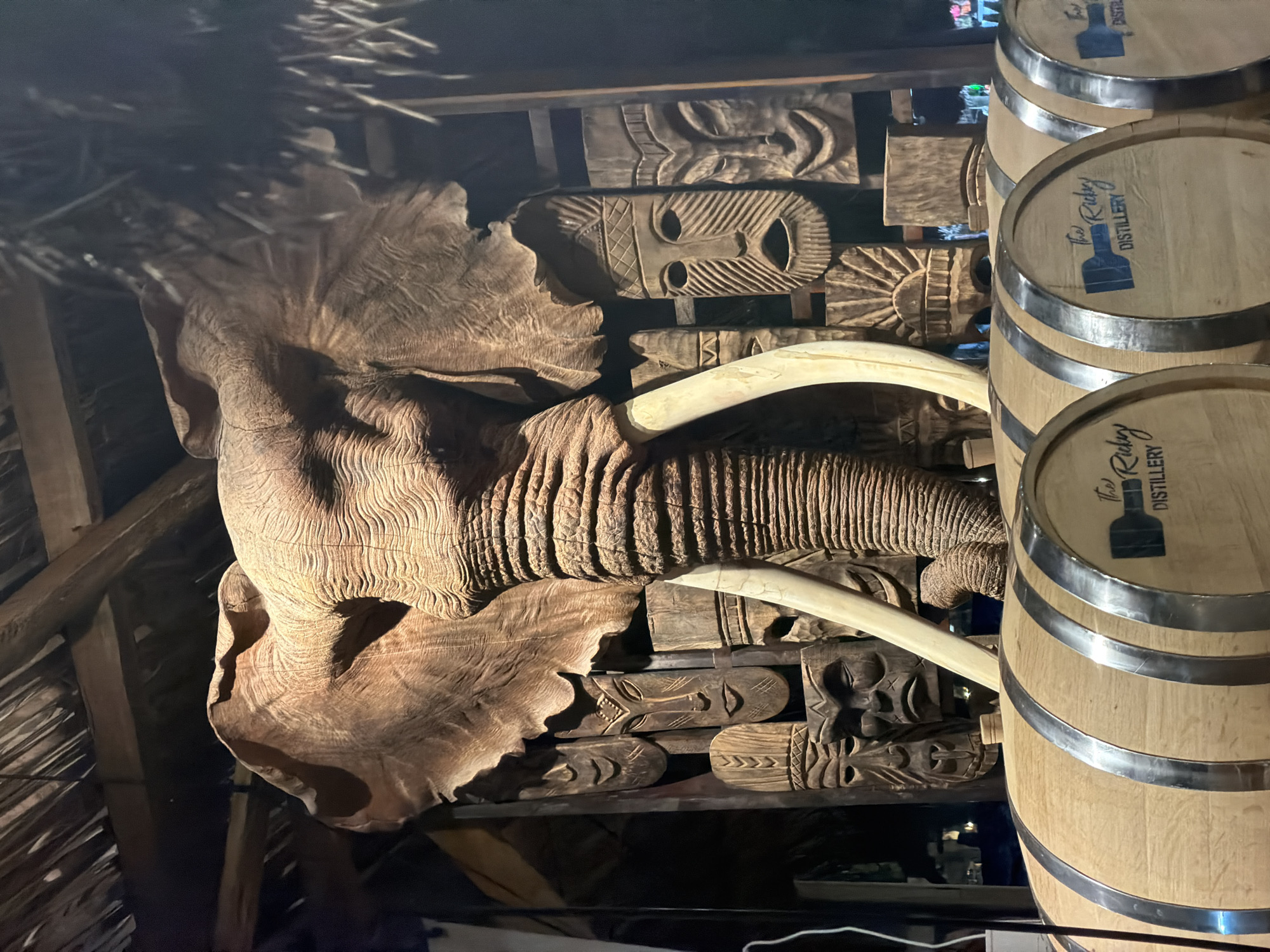 The image displays a dimly lit, rustic interior featuring a prominent, intricately carved wooden elephant head. The large carving, positioned centrally and to the left, showcases detailed textures mimicking wrinkled skin on its trunk and ears, with two long, pale tusks extending forward.
Behind and beneath the elephant head, the wall is adorned with numerous smaller wooden carvings, primarily depicting stylized tribal masks and abstract patterns, contributing to a culturally rich aesthetic. The overall environment is constructed with dark wooden beams visible in the background and a textured ceiling, possibly thatched, suggesting a traditional or themed setting.
To the right of the elephant carving, three wooden barrels are stacked vertically. Each barrel is bound with metallic hoops and features a dark logo printed on its visible end. The logo includes a silhouette of a bottle and the text "The Ridgy Distillery."
The scene is static, devoid of people or activity. The low light and strategic illumination highlight the textures and details of the wooden artwork and barrels, creating a dramatic atmosphere, indicative of an evening or indoor setting within a commercial establishment, possibly a distillery, bar, or restaurant in Măng Đen, Vietnam.