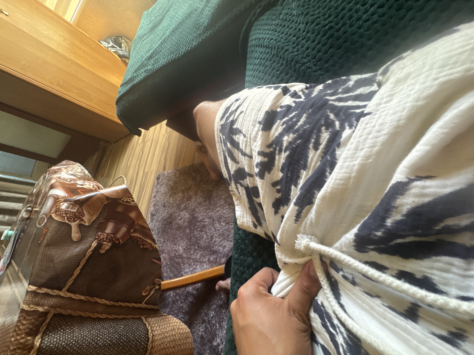 The image depicts a close-up, low-angle view of a person's legs and torso, wearing white shorts with a dark, abstract pattern. A hand is visible, holding the drawstring of the shorts. The person appears to be sitting on a dark green, textured surface, possibly a couch or chair. In the background, a round table covered with a brown tablecloth featuring coffee-themed graphics is visible. A woven basket and a piece of wood are also present. The floor appears to be wood-paneled. The lighting suggests an indoor setting, possibly during daylight hours. There is no discernible urban landscape or indication of the city of Sofia, Bulgaria.