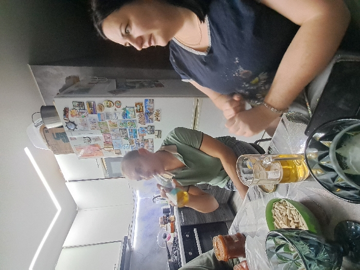 A rotated scene reveals a woman in a dark blue shirt standing behind a table laden with glasses, beer in a mug, and snacks. A man in a green shirt is present, standing near a kitchen appliance. The background includes a refrigerator covered in magnets and papers, and a kitchen with white cabinets.