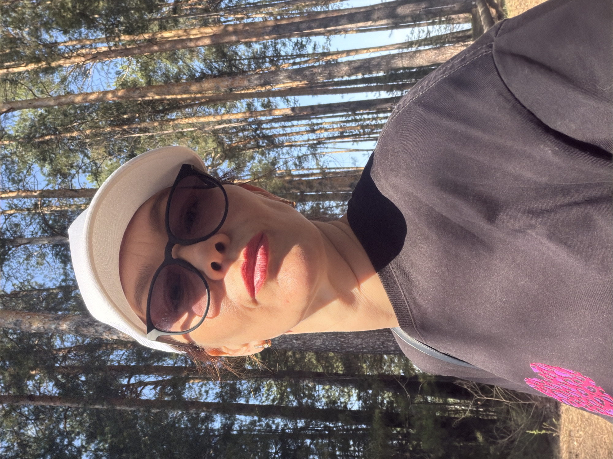 The photograph shows a woman in an outdoor setting with a backdrop of tall trees. She is wearing a black t-shirt with a pink design on the shoulder, a white hat, and sunglasses. Her red lipstick stands out. The trees behind her have bare branches, suggesting it could be late fall or early spring. Light filters through the trees, creating a dappled effect. The location is in a wooded area in the city of Chaikovsky, Russia.