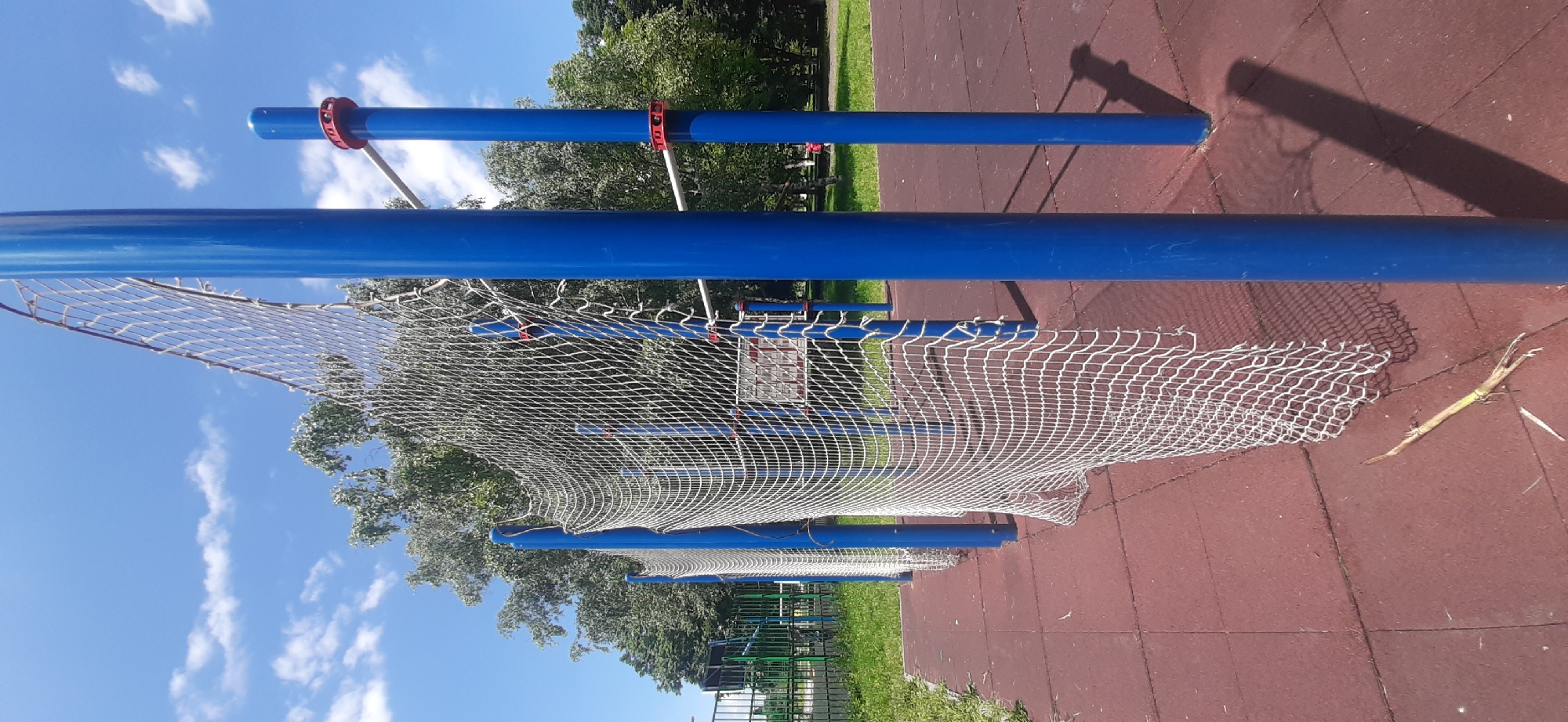 A sports court in Yessentukskaya, Russia, features a net supported by tall, blue poles. The net, made of white mesh, is partially obscured by the poles and extends into the background. The court is surfaced with red, rubberized tiles. Green grass and trees are visible beyond the court, under a bright blue sky with scattered white clouds.