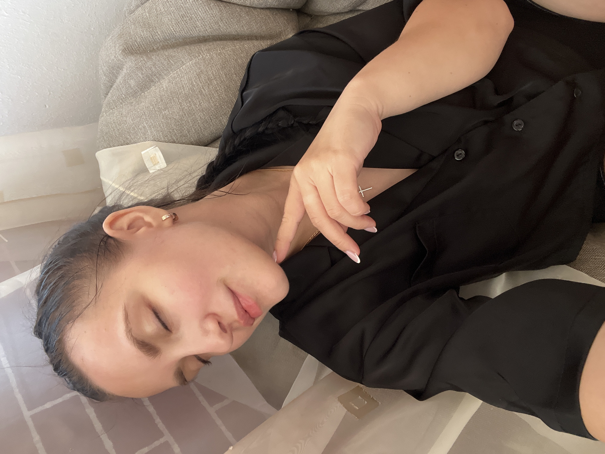 The image shows a young woman resting or sleeping indoors, lying on her side. Her head is positioned on a light-colored surface, possibly a pillow or blanket with a faint grid pattern, next to a beige, textured couch or headboard and a white wall. Her eyes are closed, and her facial expression appears peaceful and relaxed.
She has dark hair, which looks slightly damp or pulled back from her face. She is wearing a black collared shirt, unbuttoned at the top. Around her neck, a gold chain necklace with a small cross pendant is visible. A small gold hoop earring can be seen in her right ear. Her right hand is gently placed near her chest, revealing manicured nails with a French tip design.
The lighting is soft and ambient, characteristic of an indoor setting. On a small white tag or label visible near her head, the partial text "EGO." can be discerned. The scene conveys a sense of calm and repose. The provided location context for this media file is Марковский, Russia.
