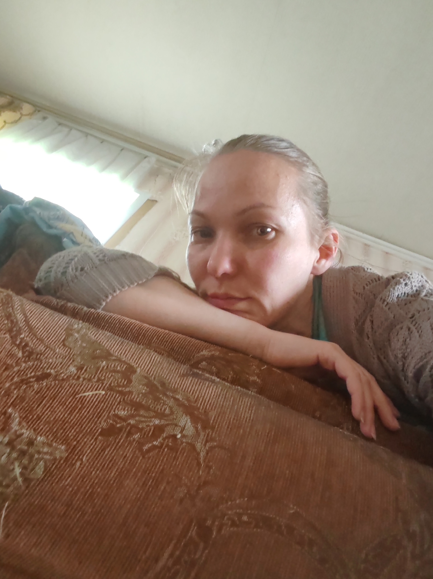 A woman is depicted in a close-up, low-angle shot, looking directly at the camera. She has light-colored hair pulled back and appears to be resting her left arm and elbow on a large piece of brown, patterned upholstered furniture, possibly a couch or bed. She is wearing a light brown or grey knitted cardigan over a teal or green top. Her expression is neutral, possibly pensive or tired.
The brown furniture fills the lower and mid-left portions of the frame, showing a textured, ribbed fabric with a subtle darker pattern. In the background, to the left of the woman's head, there is a brightly lit window with light-colored blinds or curtains, suggesting daytime. Above the window and extending across the upper right, a plain, light-colored wall or ceiling is visible. A dark, possibly folded garment or fabric is visible on the brown furniture near the woman's left shoulder. The scene takes place indoors, in what appears to be a domestic setting. There is no visible text in the image. The location context provided is Чухлэм, Russia.