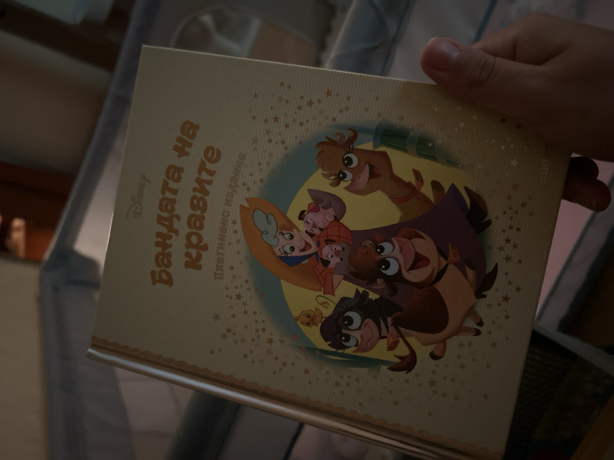 The image shows a person holding a children's book with a light yellow cover. The book has a colorful illustration of cartoon characters, including what appears to be a woman in a purple cape and several animal characters such as a piglet, a cow, a llama, and a monkey. There are also small star patterns scattered across the cover. The title "Бандата на кравите" (The Cow Gang) is visible in orange lettering at the top, with "Disney" written above it. Below the title, "Платинено издание" (Platinum Edition) is printed. The background suggests an indoor setting, possibly a nursery or child's room, with parts of a crib visible. The lighting is dim, and the focus is on the book. There is no discernible location in the city of Sofia, Bulgaria, specified in the image.