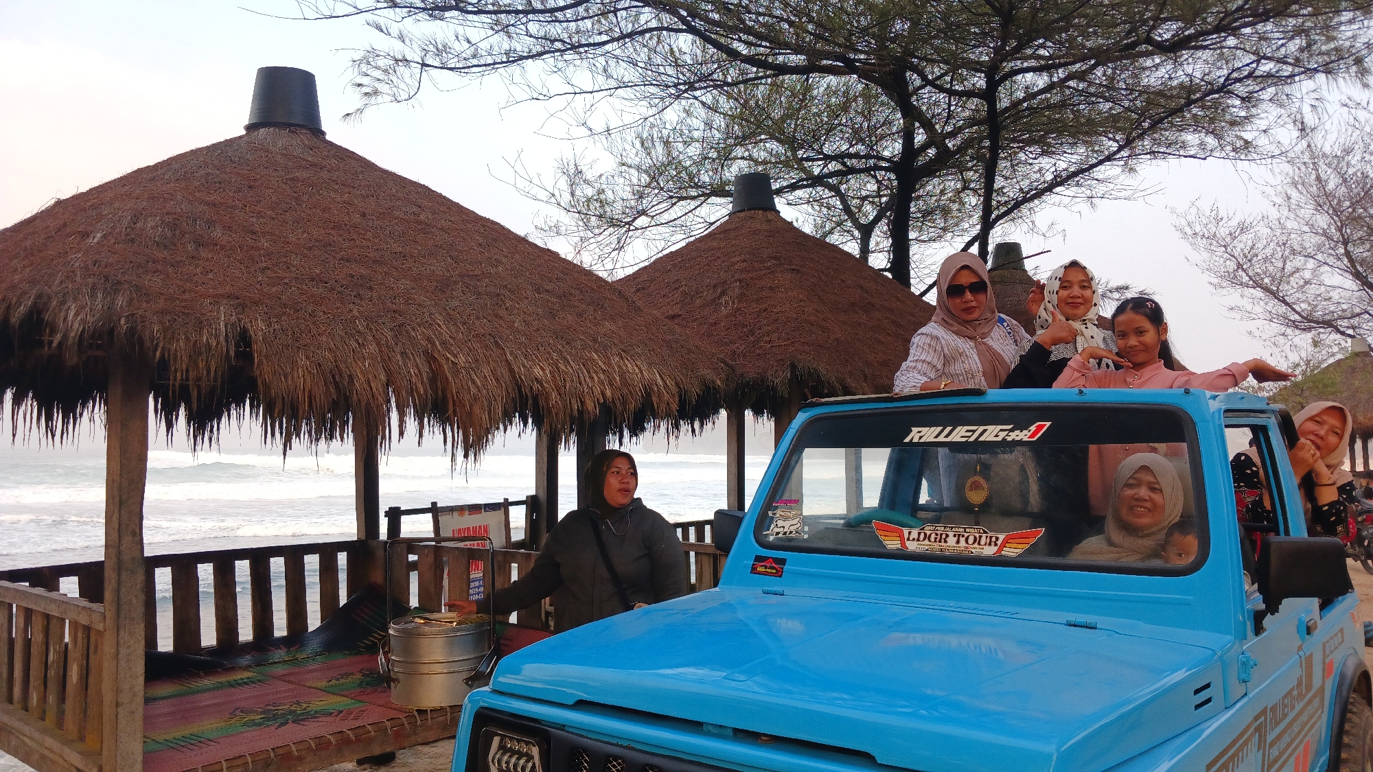 The image shows a group of people at a beach in Tepus, Indonesia. In the foreground, a bright blue jeep is parked, with several people posing on top and inside it. One woman in a hijab is leaning out of the driver's side window, smiling. Another woman in a grey hijab is seated in the passenger seat, looking at the camera. Three women and a young girl are standing on top of the jeep, with the young girl in the center striking a pose. Behind the jeep, there are two thatched-roof huts, typical of beachside establishments. The huts have wooden railings and what appears to be seating inside. The ocean is visible in the background, with waves crashing on the shore. The weather appears to be overcast. A woman in a dark jacket is standing next to the huts, facing away from the jeep. There is also a metal steaming pot on a mat inside one of the huts. The scene depicts a casual outing at a coastal location.