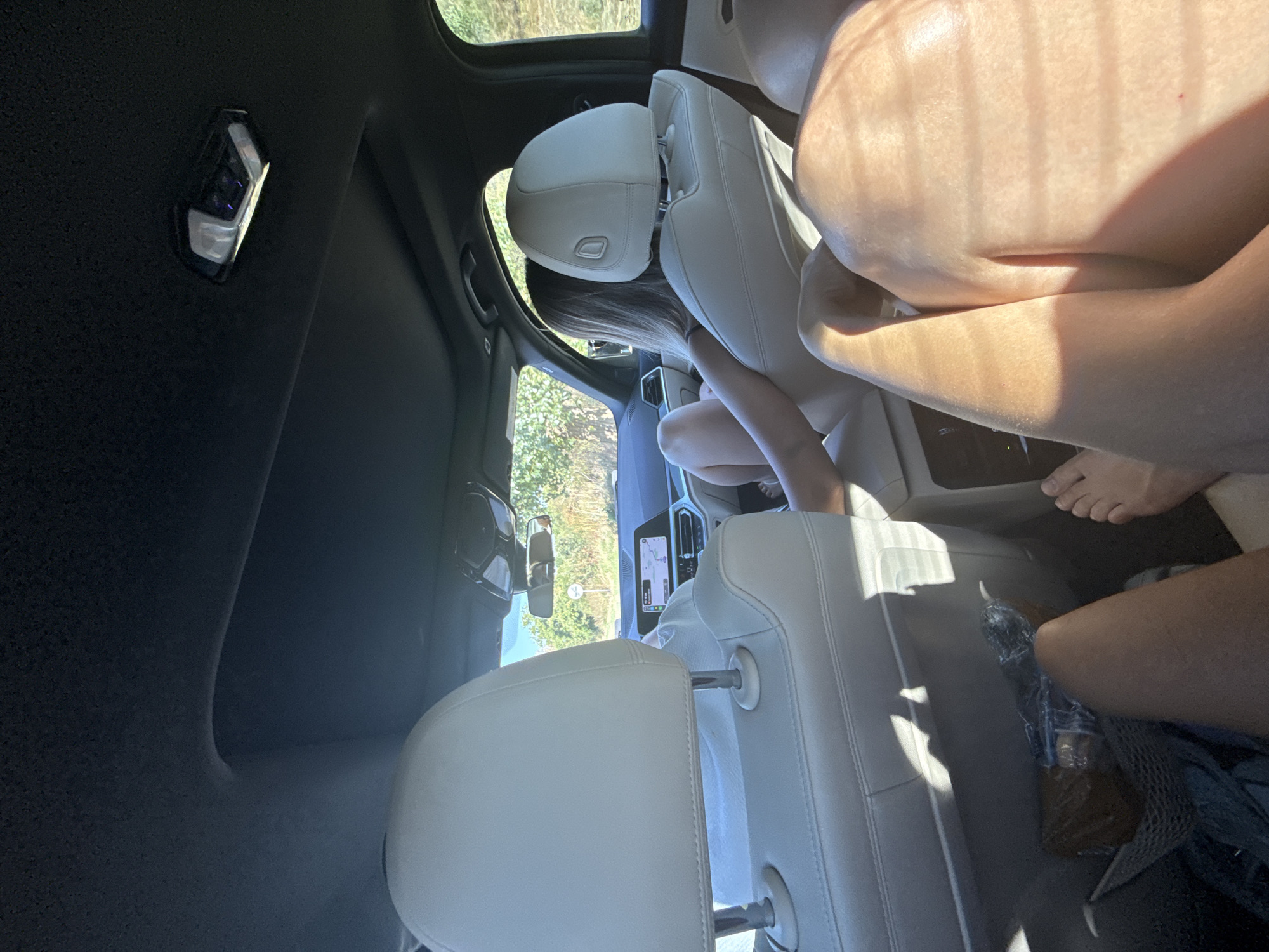 The image shows the interior of a car from the perspective of a passenger in the back seat. The rear headrests of a light-colored leather interior are prominently displayed in the foreground. A person, mostly obscured, is seated in the front passenger seat, their legs visible. Another person, with blonde hair, is seated in the rear driver's side seat, their body angled away from the camera. They are wearing a light-colored top. The car's dashboard and infotainment screen are visible through the front seats, displaying a navigation map. Outside the car, through the windows, a sunny, rural landscape with green foliage and trees can be seen, suggesting a daytime journey on a paved road. The sky is clear and blue. The image does not contain any specific landmarks or clues that would allow for precise identification of the location within Căbești, Romania. The scene depicts a typical car interior with passengers on a road trip.