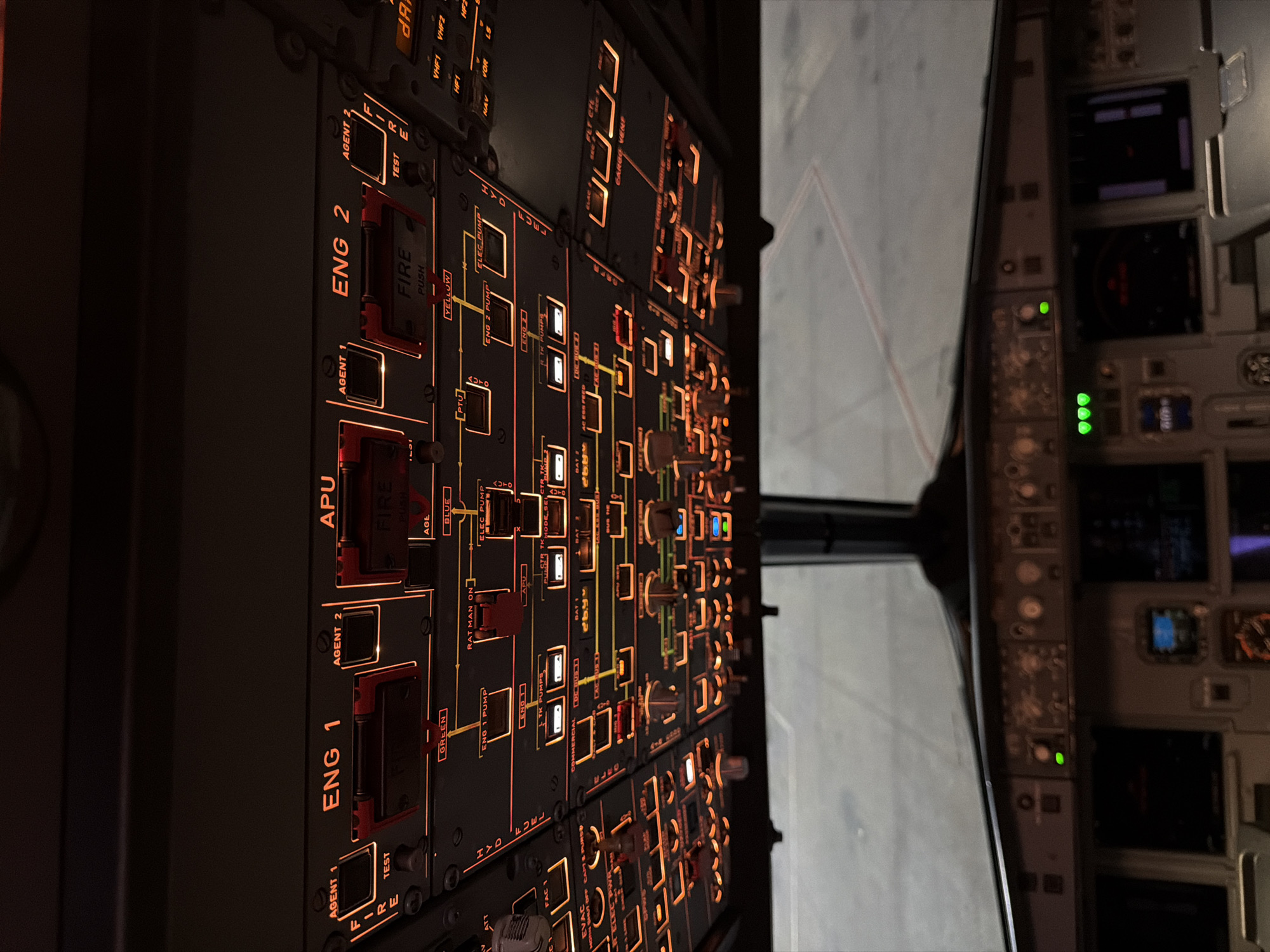 The image shows the cockpit of an airplane. The control panel is the main focus, with numerous buttons and switches illuminated in orange. Some key labels visible include "ENG 1", "ENG 2", "APU", "FIRE", "FUEL", and "HYD". The windshield and the view outside the plane can also be seen. The exterior view appears to be a tarmac or runway with a light-colored concrete surface.